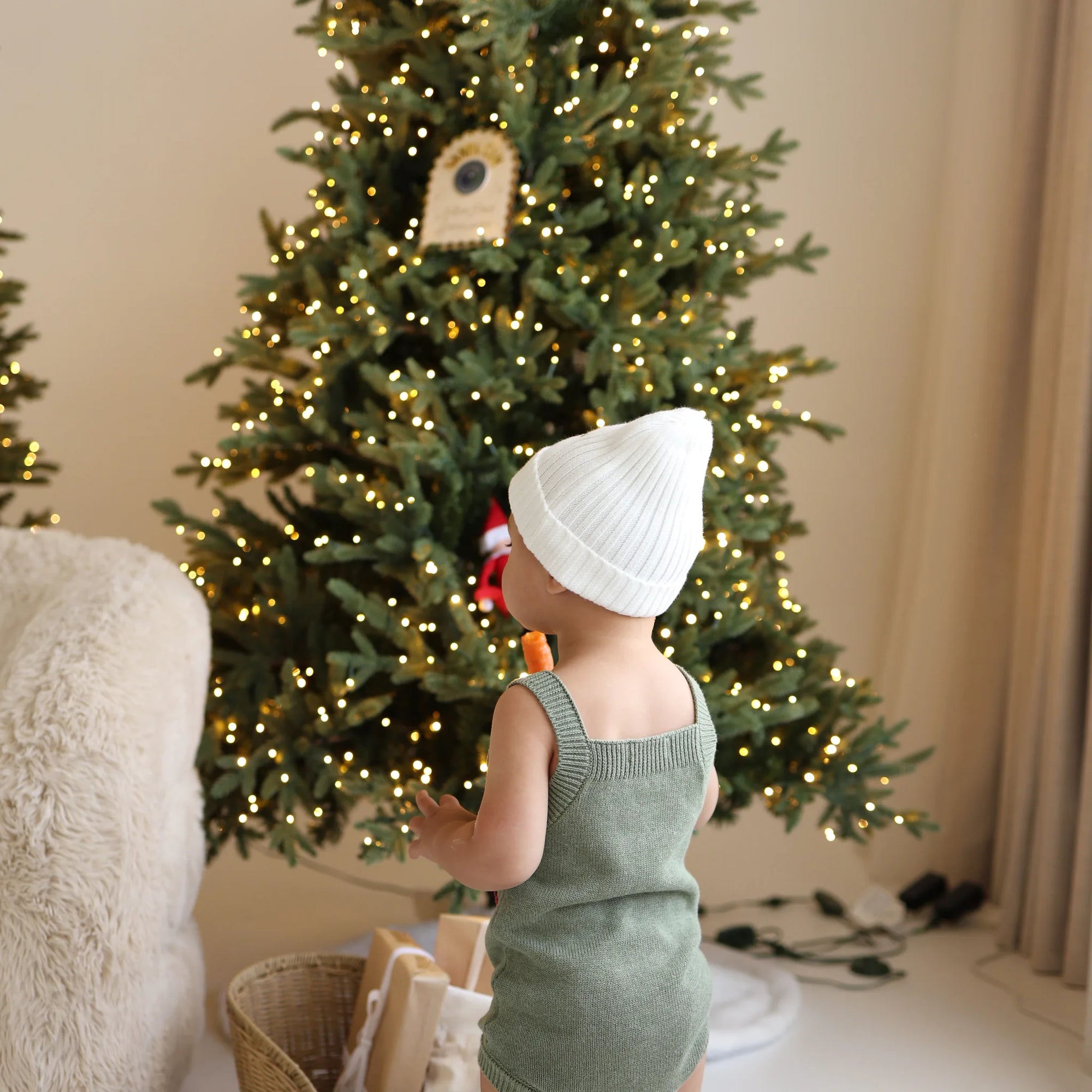 Back view of a baby in a green knit Merry romper and white beanie looking up at a lit Christmas tree
