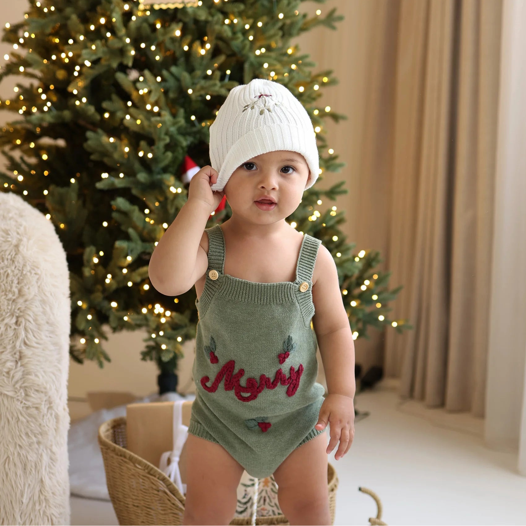 Baby in a green knit Merry romper reaching up to a white ribbed beanie, lit Christmas trees behind