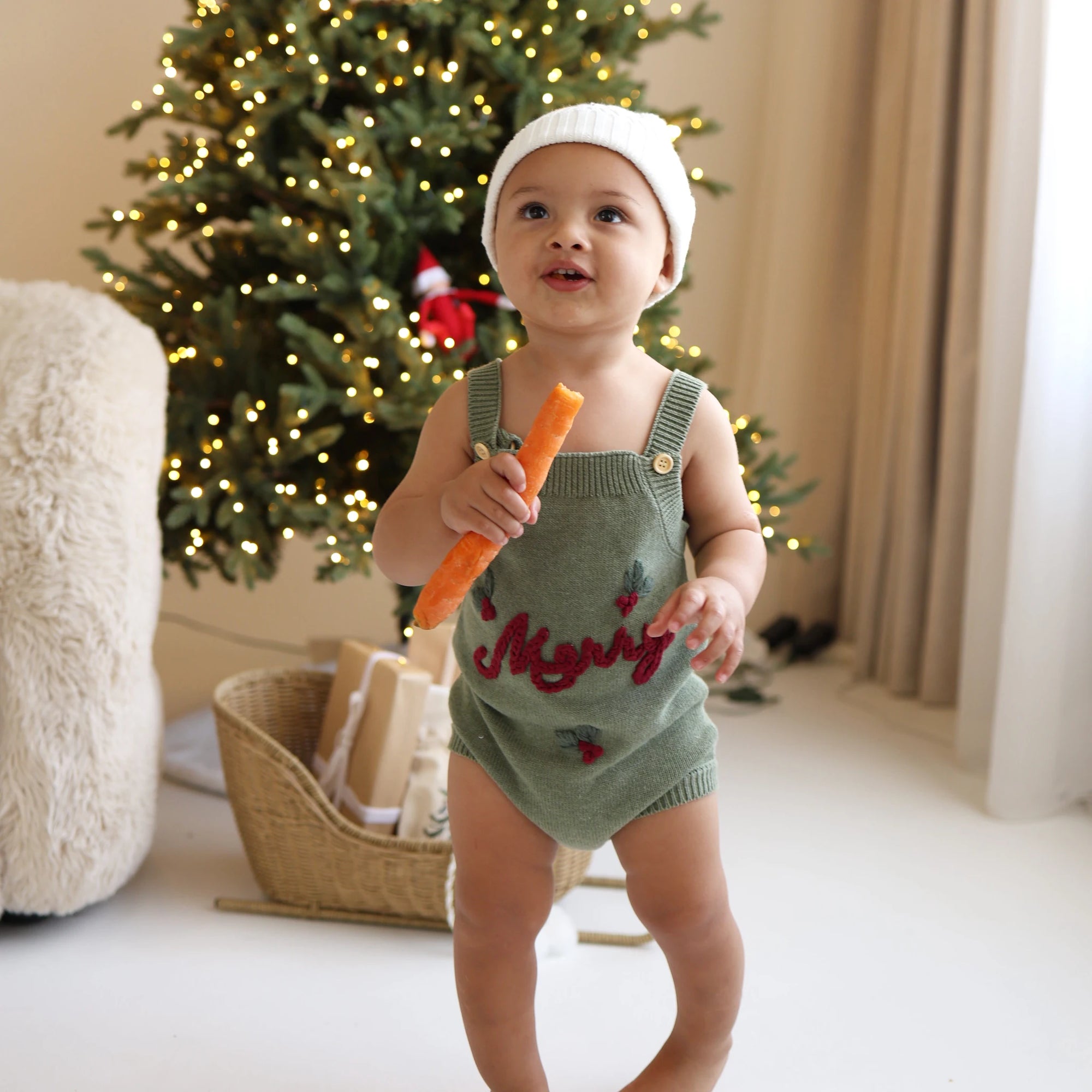 Baby in a green knit romper embroidered with Merry and a white beanie, holding a carrot for the reindeer