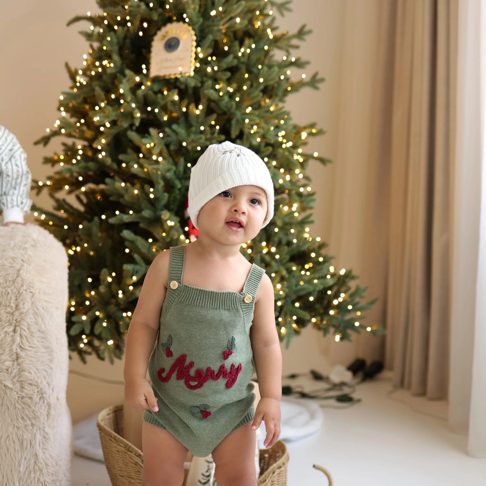 Baby in a green knit overall romper embroidered with Merry and a white ribbed beanie, standing in front of a lit Christmas tree
