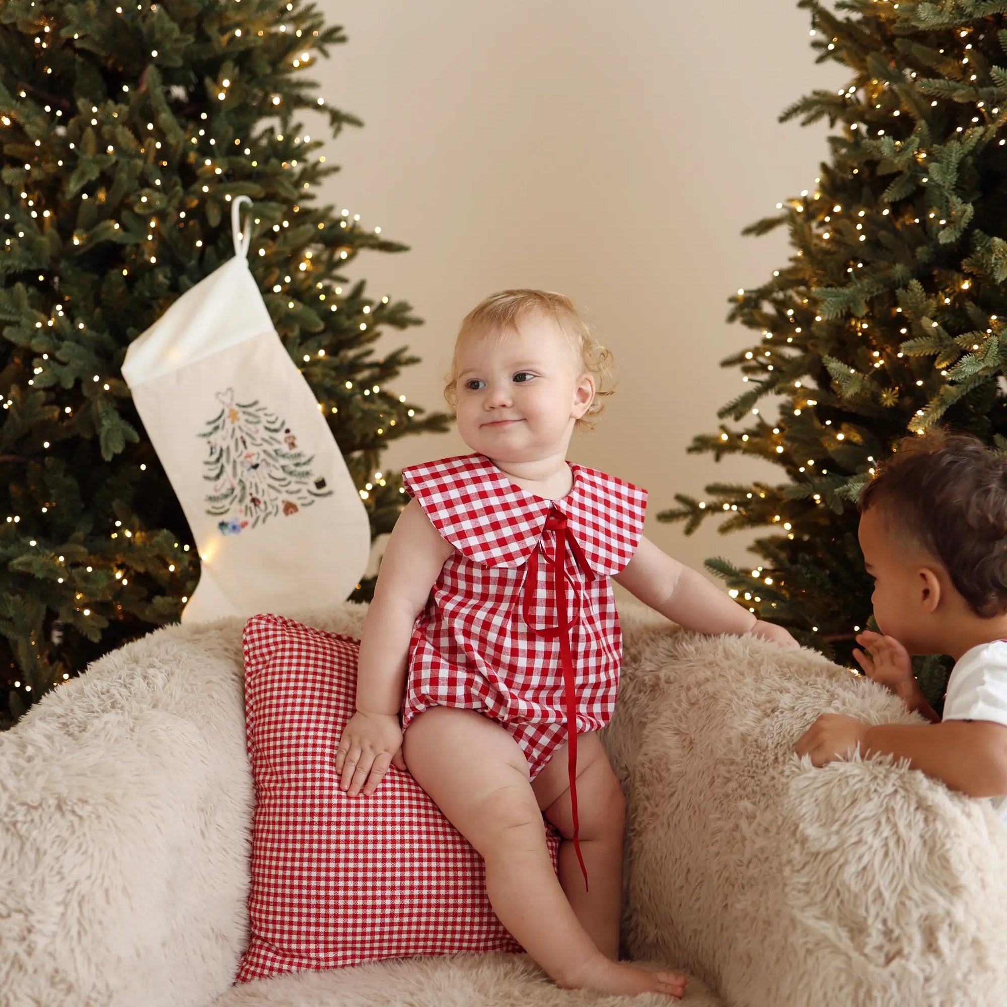 Baby in a red gingham collared romper sitting on a sheepskin chair between lit Christmas trees