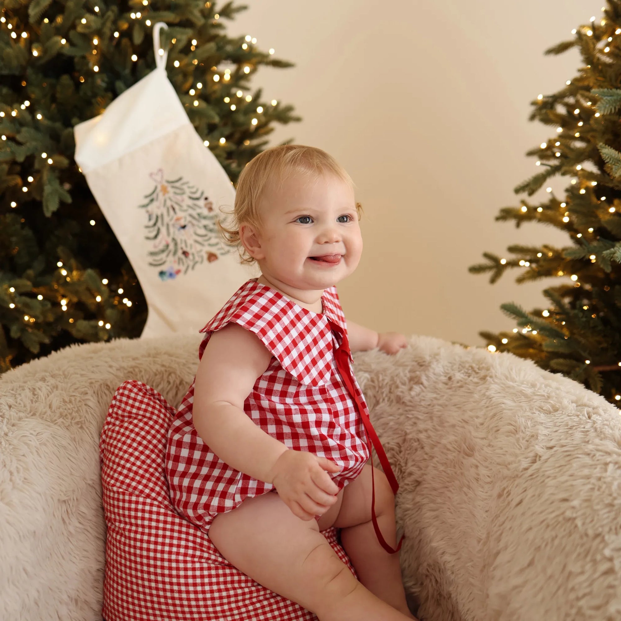 Smiling baby in a red gingham collared romper on a sheepskin chair beside an embroidered stocking