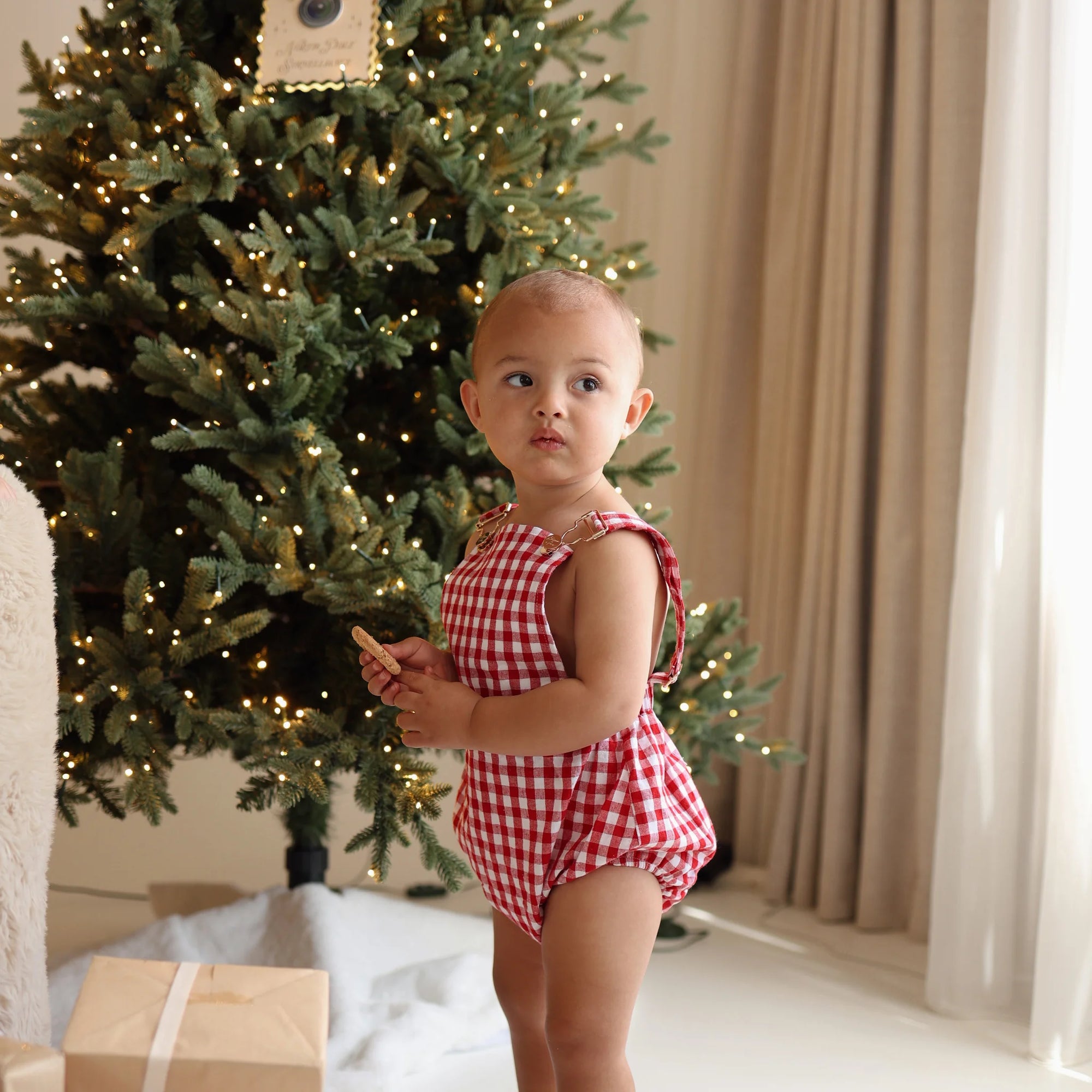 Baby in a red gingham tie shoulder romper holding a cookie in front of a lit Christmas tree