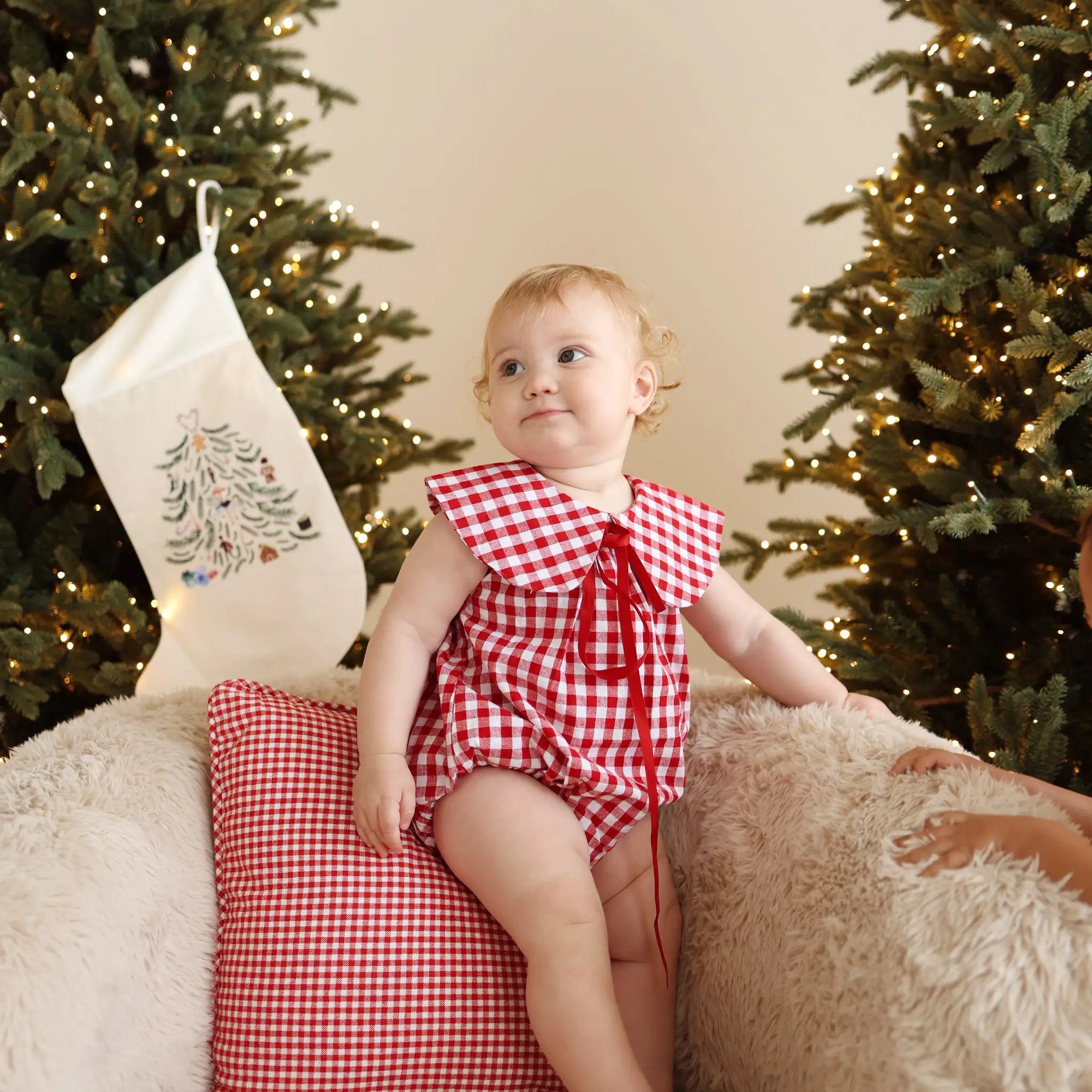 Baby in a red gingham romper with an oversized collar and red tie, on a sheepskin chair by a lit tree