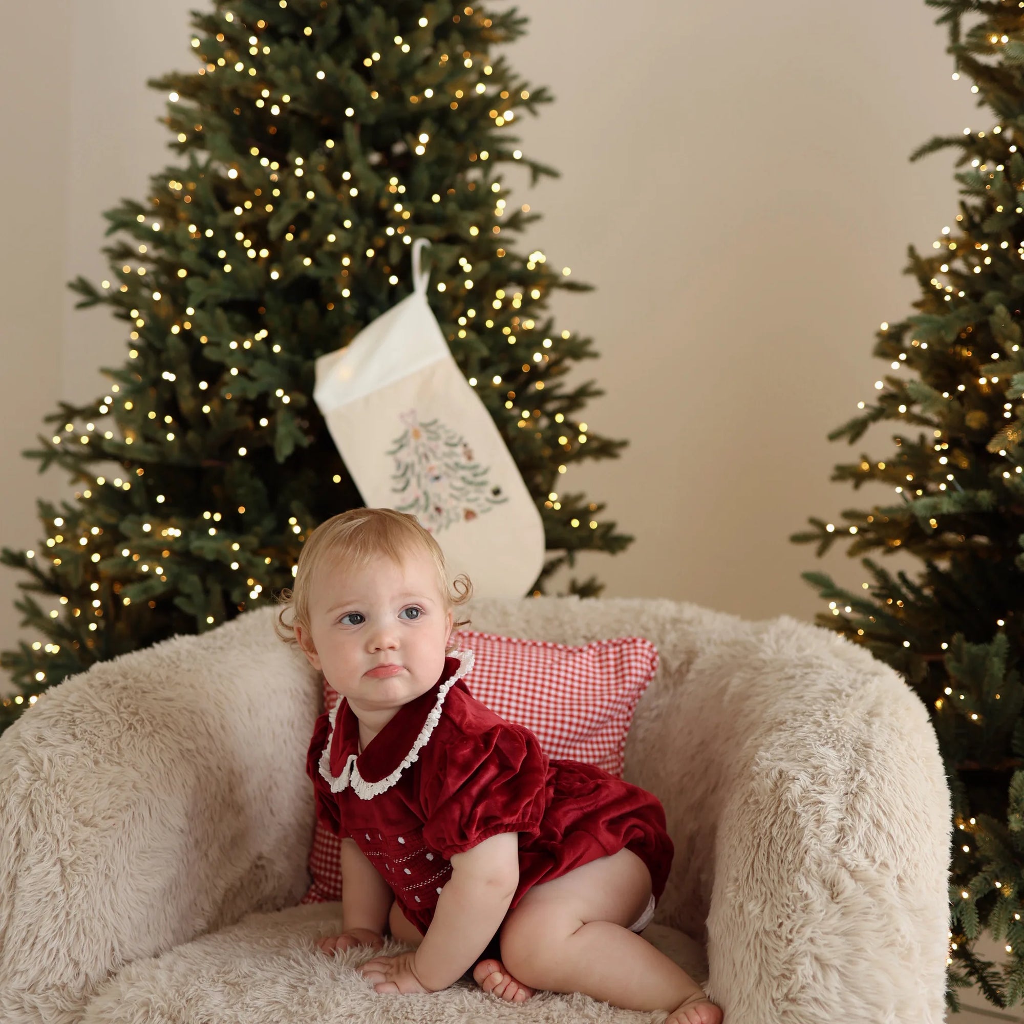 Baby in a red velvet Christmas dress with lace collar sitting on a sheepskin chair between lit trees