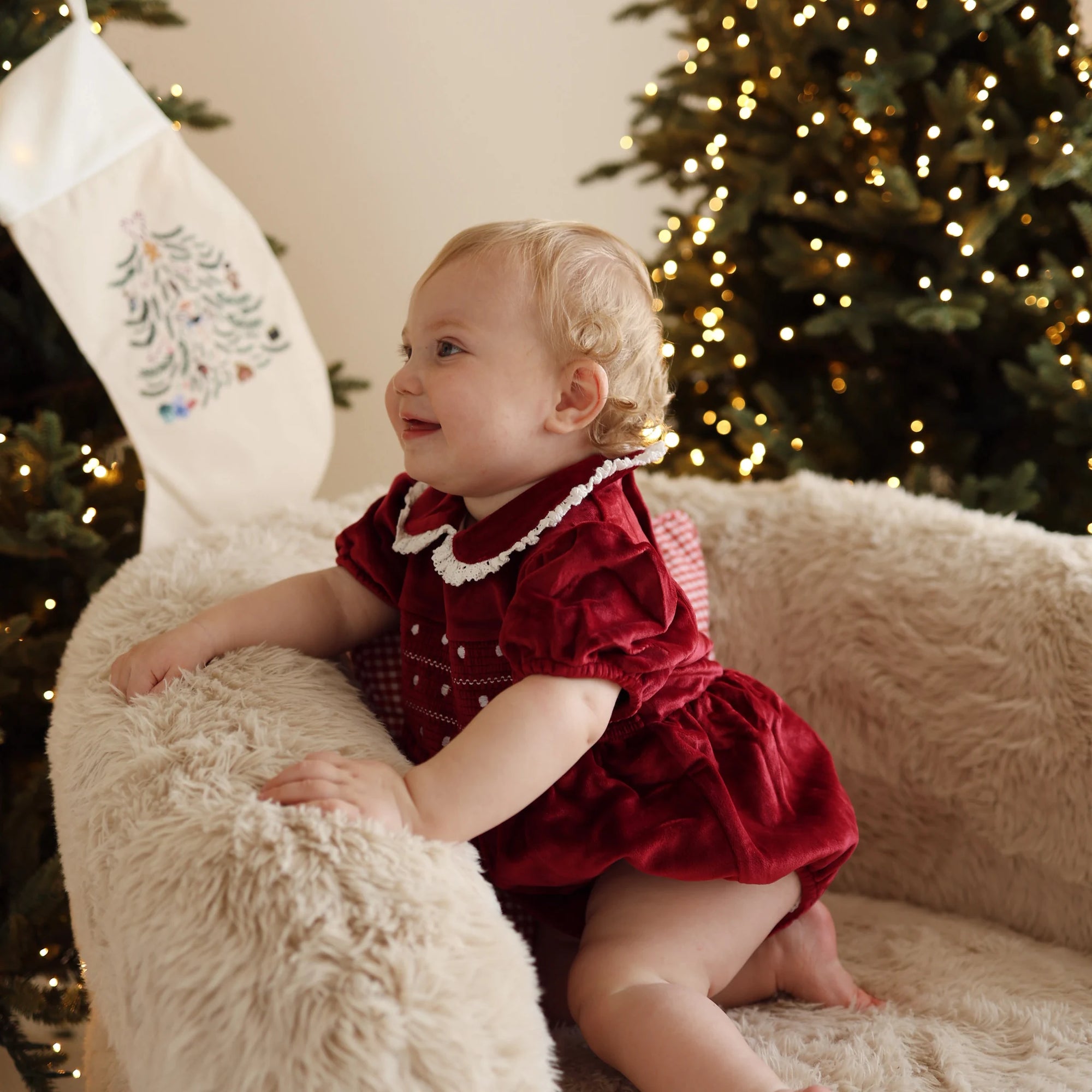 Baby in a red velvet Christmas dress with lace collar on a sheepskin chair beside an embroidered stocking