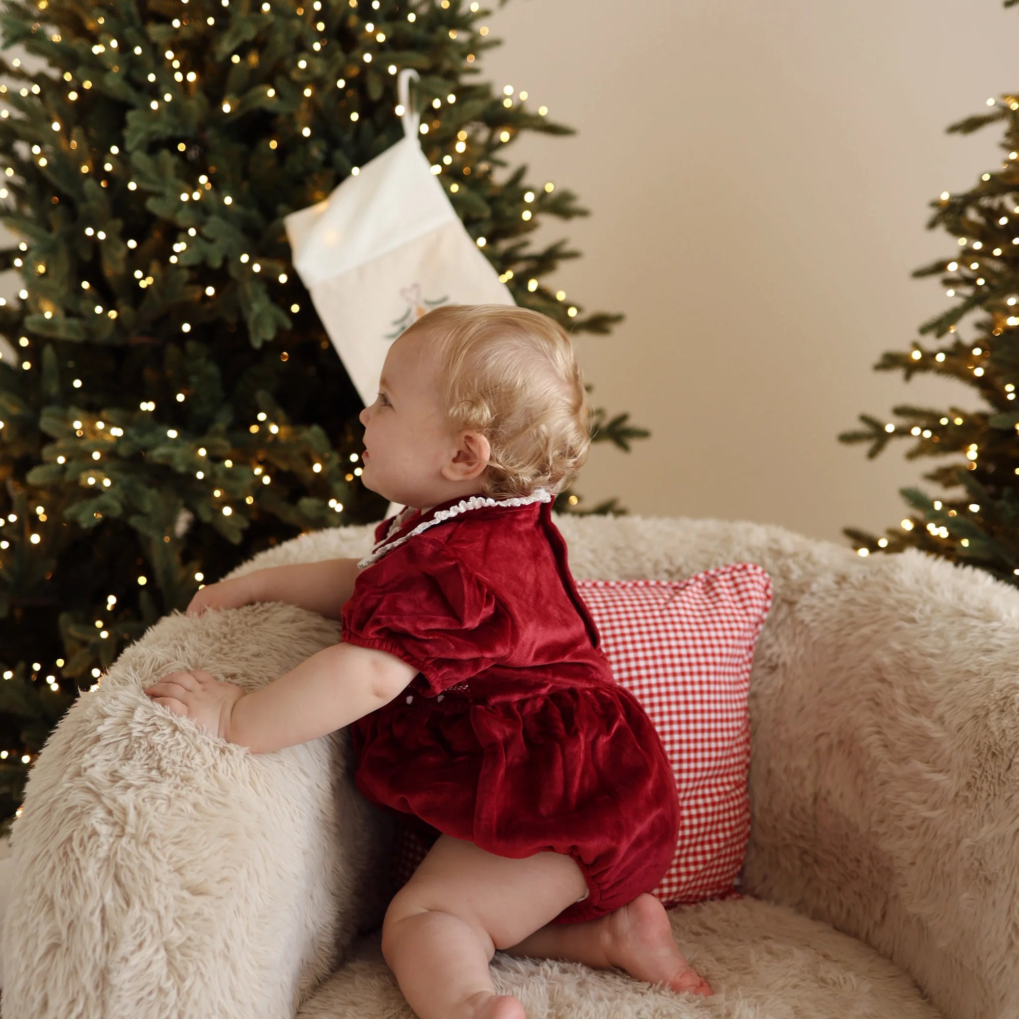 Baby in a red velvet romper with lace collar looking up at an embroidered Christmas stocking