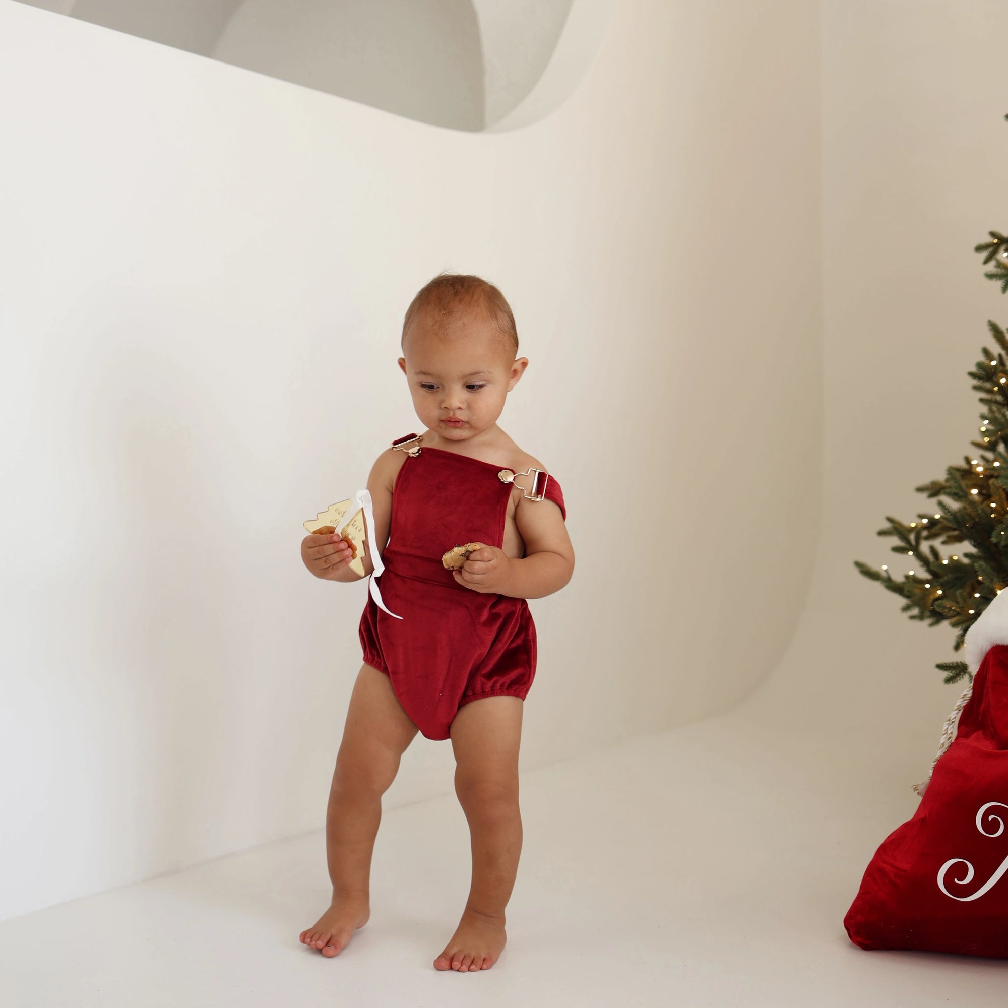Baby in a red velvet Christmas romper walking beside a personalised red Santa sack