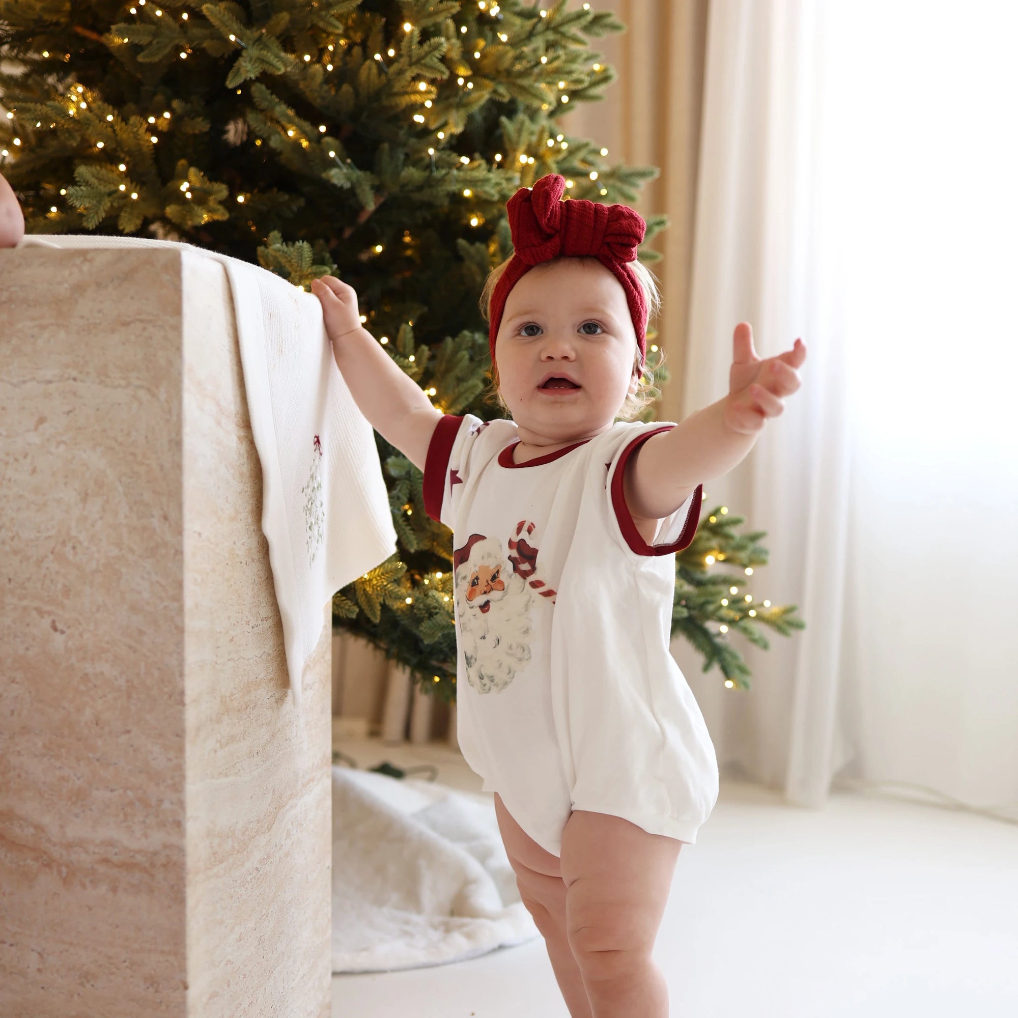 Baby in a vintage Santa print romper and red knit bow headband reaching up beside a lit Christmas tree