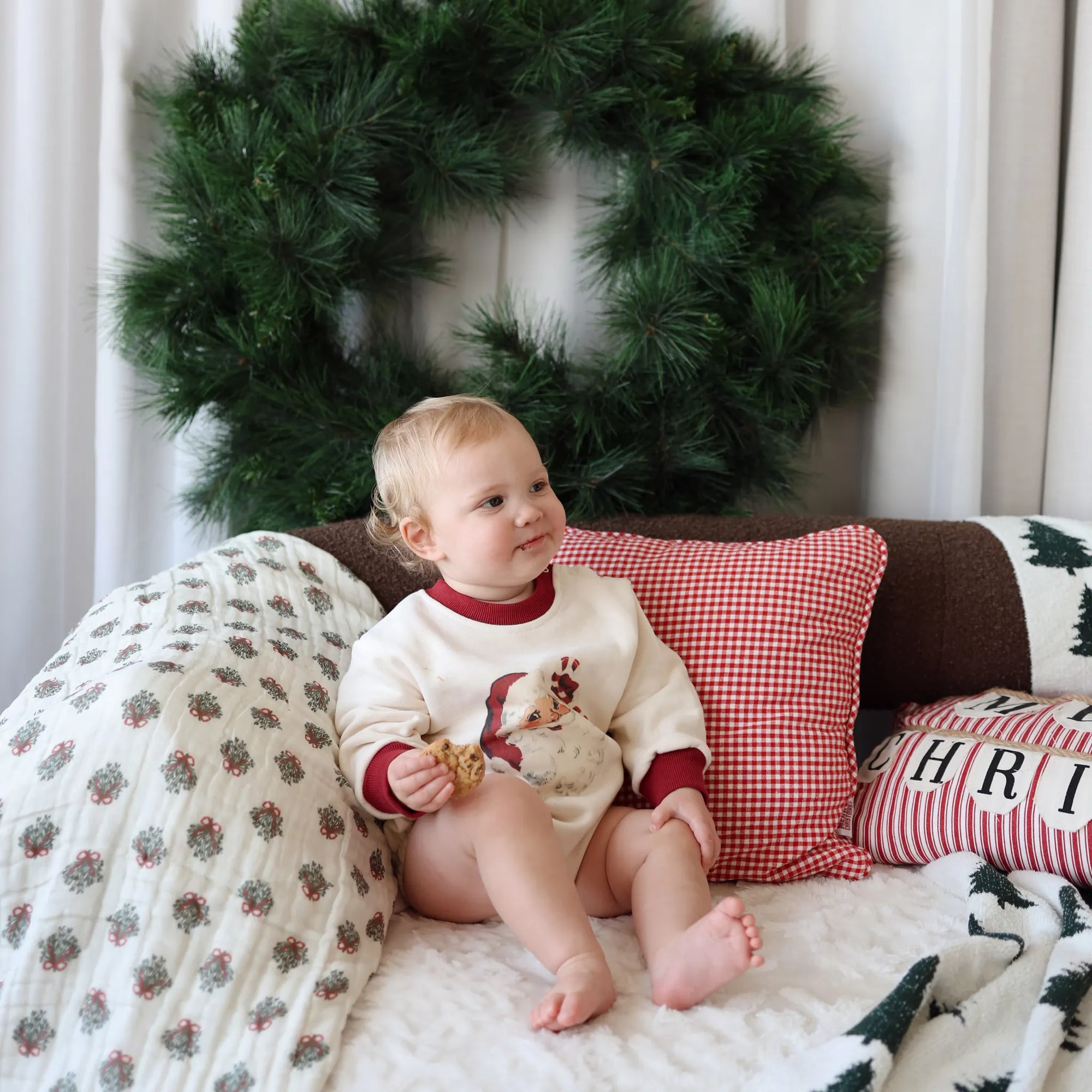 Baby in a vintage Santa print romper sitting on a sofa with Christmas cushions and a wreath behind