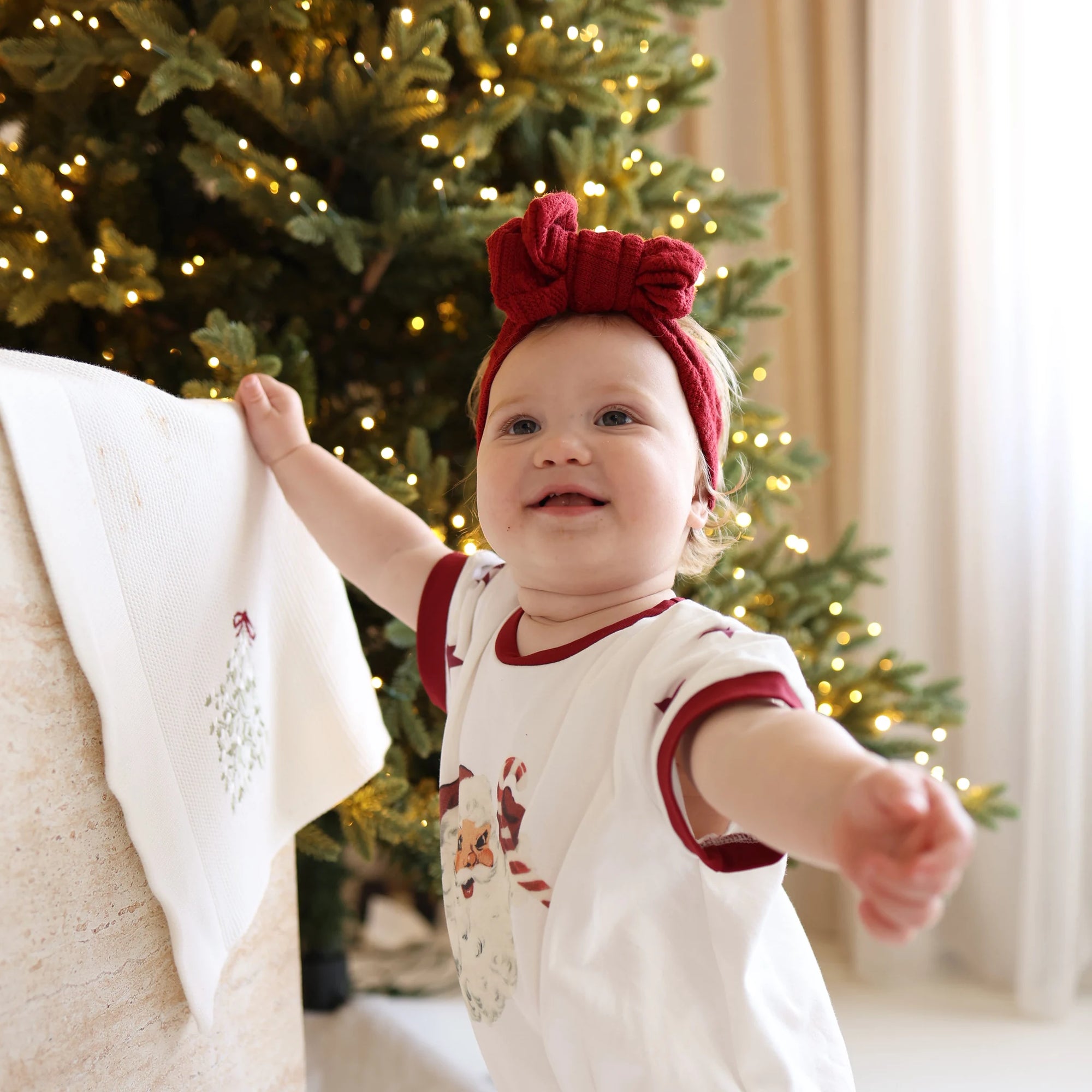 Baby in a Santa print tee set and red knit bow headband reaching for an embroidered mistletoe stocking