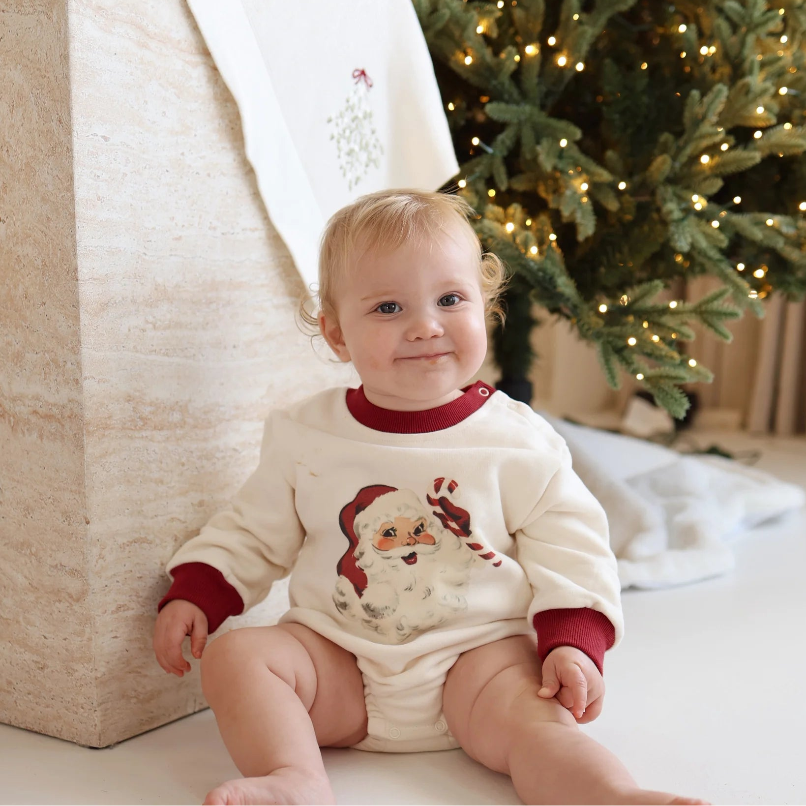 Baby in a cream vintage Santa print romper with red cuffs, sitting in front of a lit Christmas tree