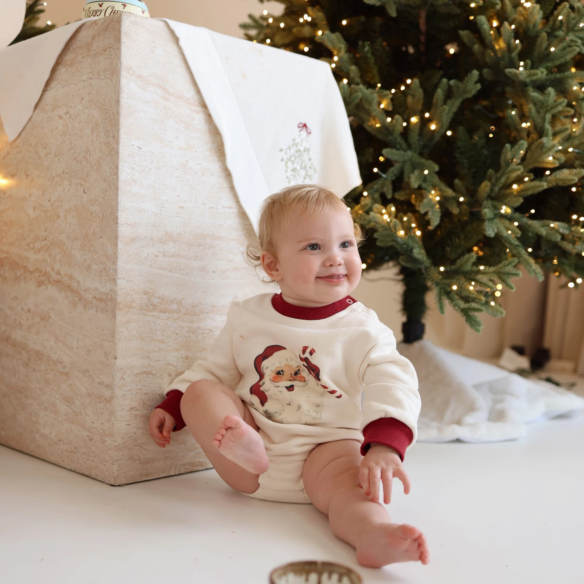 Smiling baby in a cream vintage Santa print romper with red trim, sitting beside a lit Christmas tree