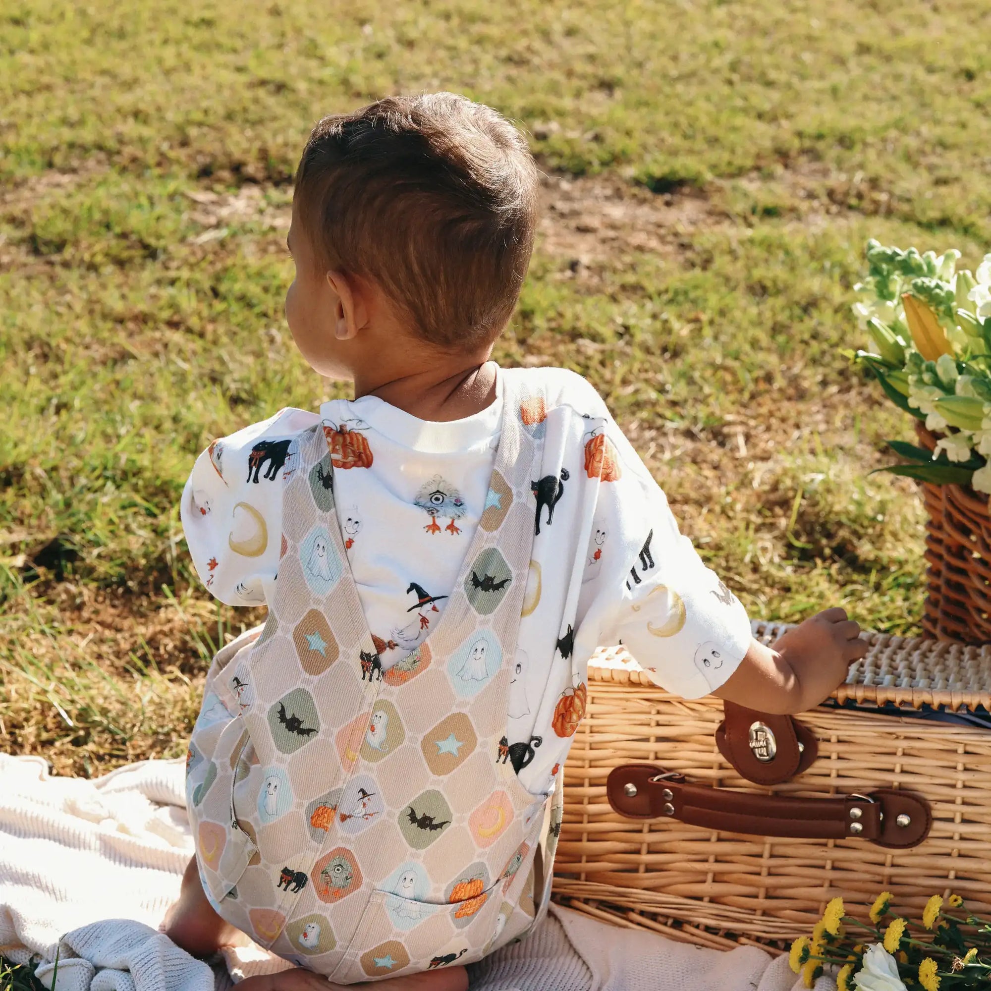 Back view of a toddler in Halloween print overalls and a matching print shirt sitting beside a picnic basket