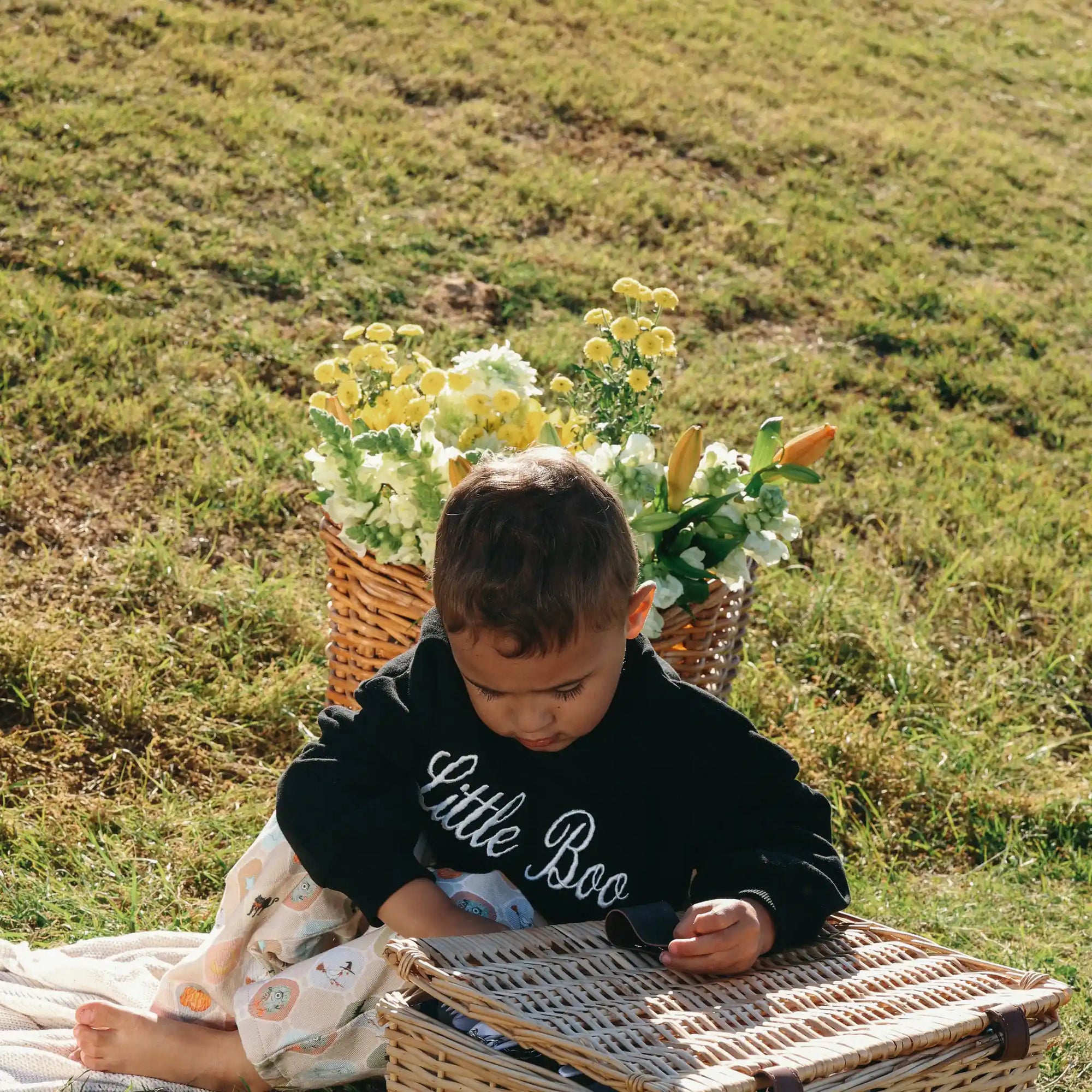 Toddler in a black Little Boo embroidered sweatshirt opening a picnic basket beside flowers on grass