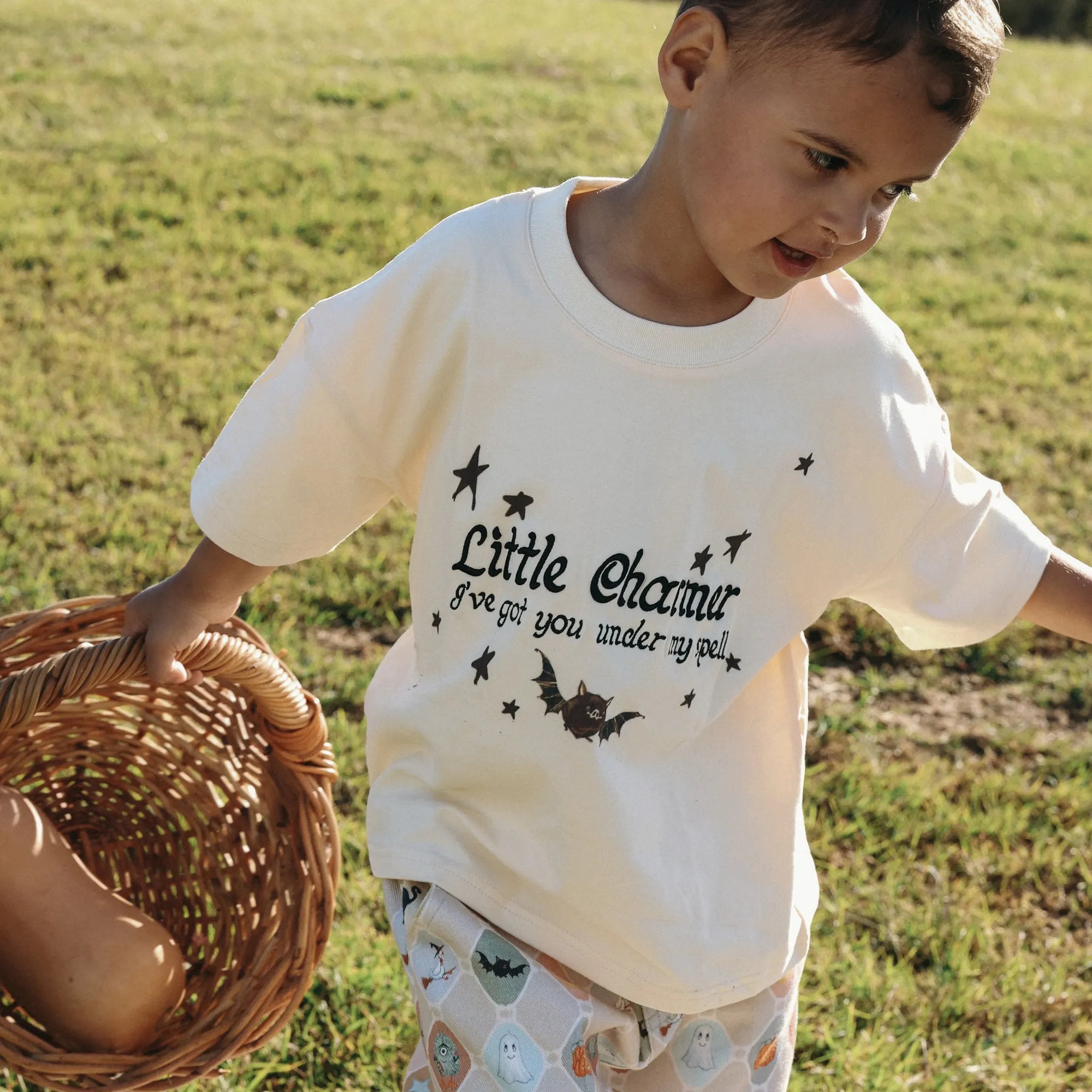 Toddler in a Little Charmer I've Got You Under My Spell tee lifting a wicker basket in a field