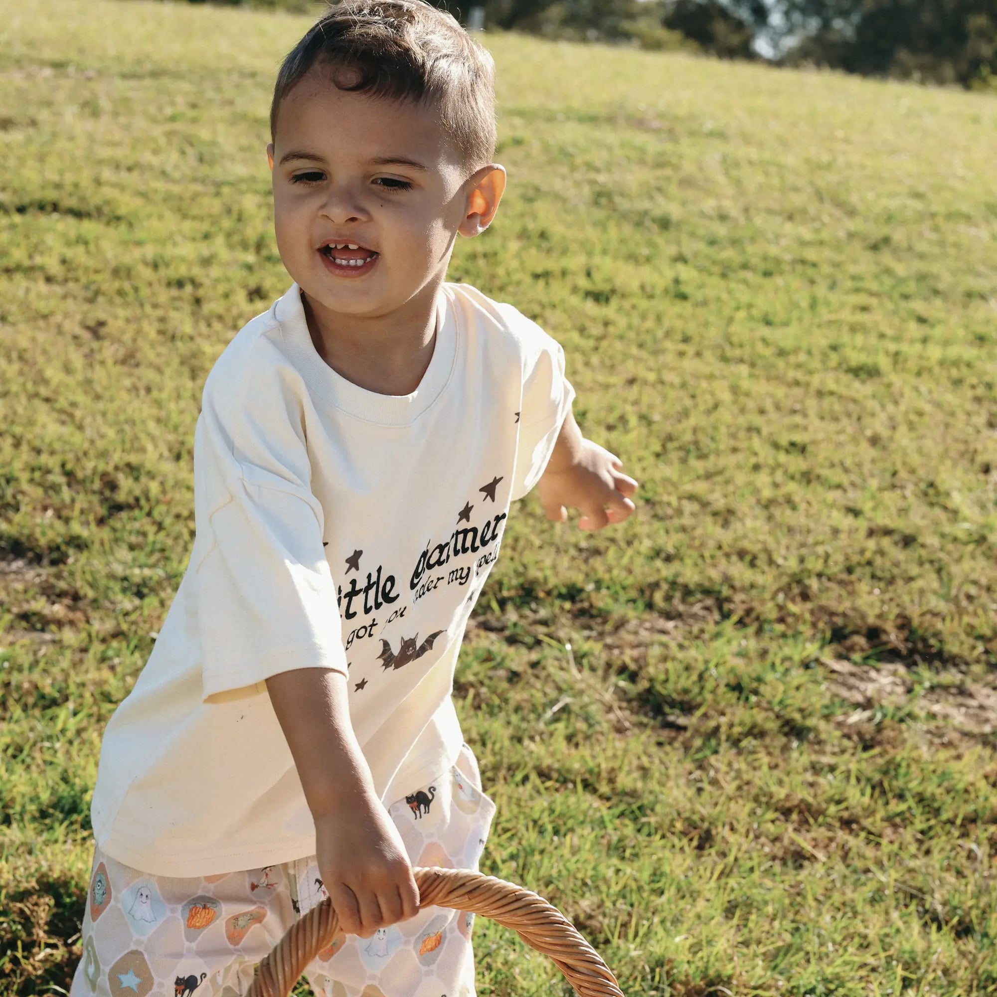 Smiling toddler in a Little Charmer tee and Halloween print pants holding a wicker basket handle