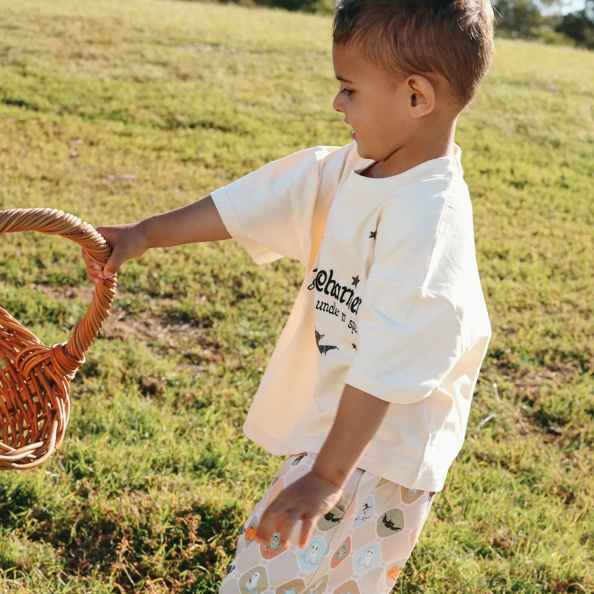 Toddler in a Little Charmer tee and Halloween print pants pulling a wicker basket across the grass