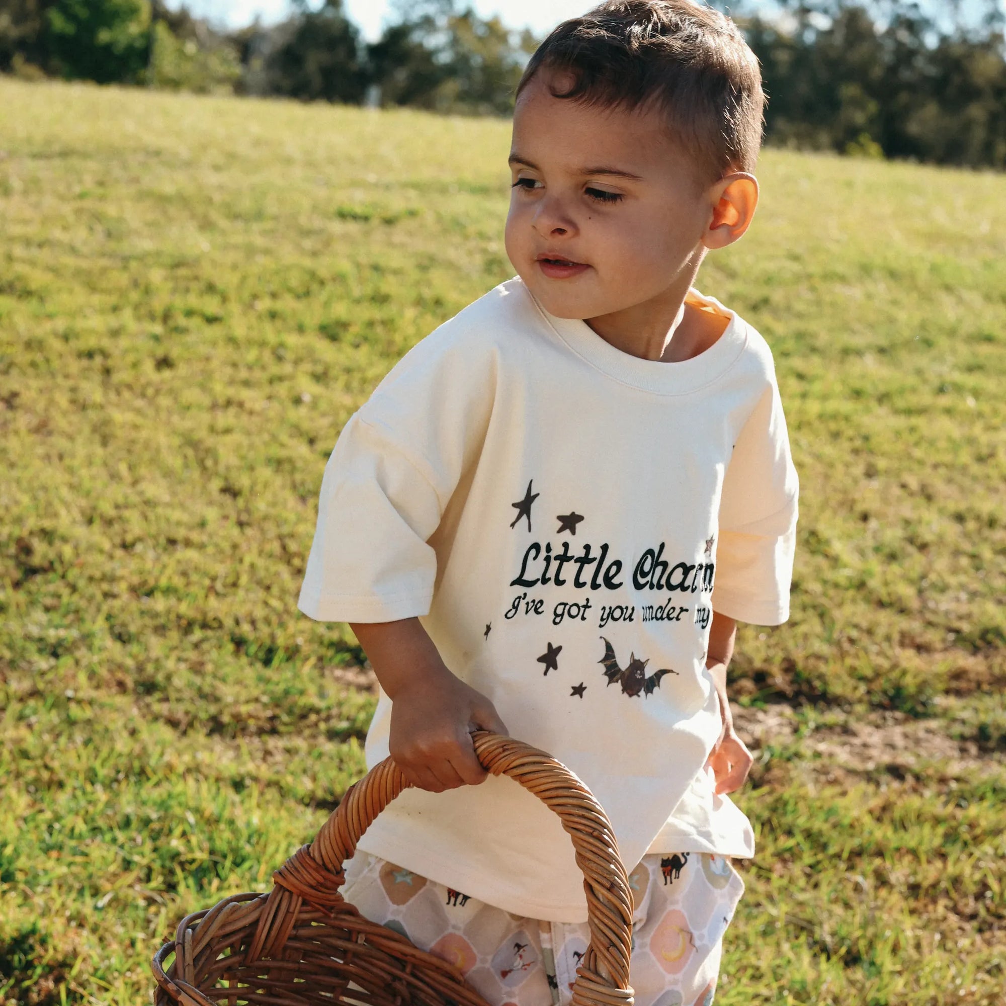 Toddler in a Little Charmer tee and Halloween print pants carrying a wicker basket across the grass