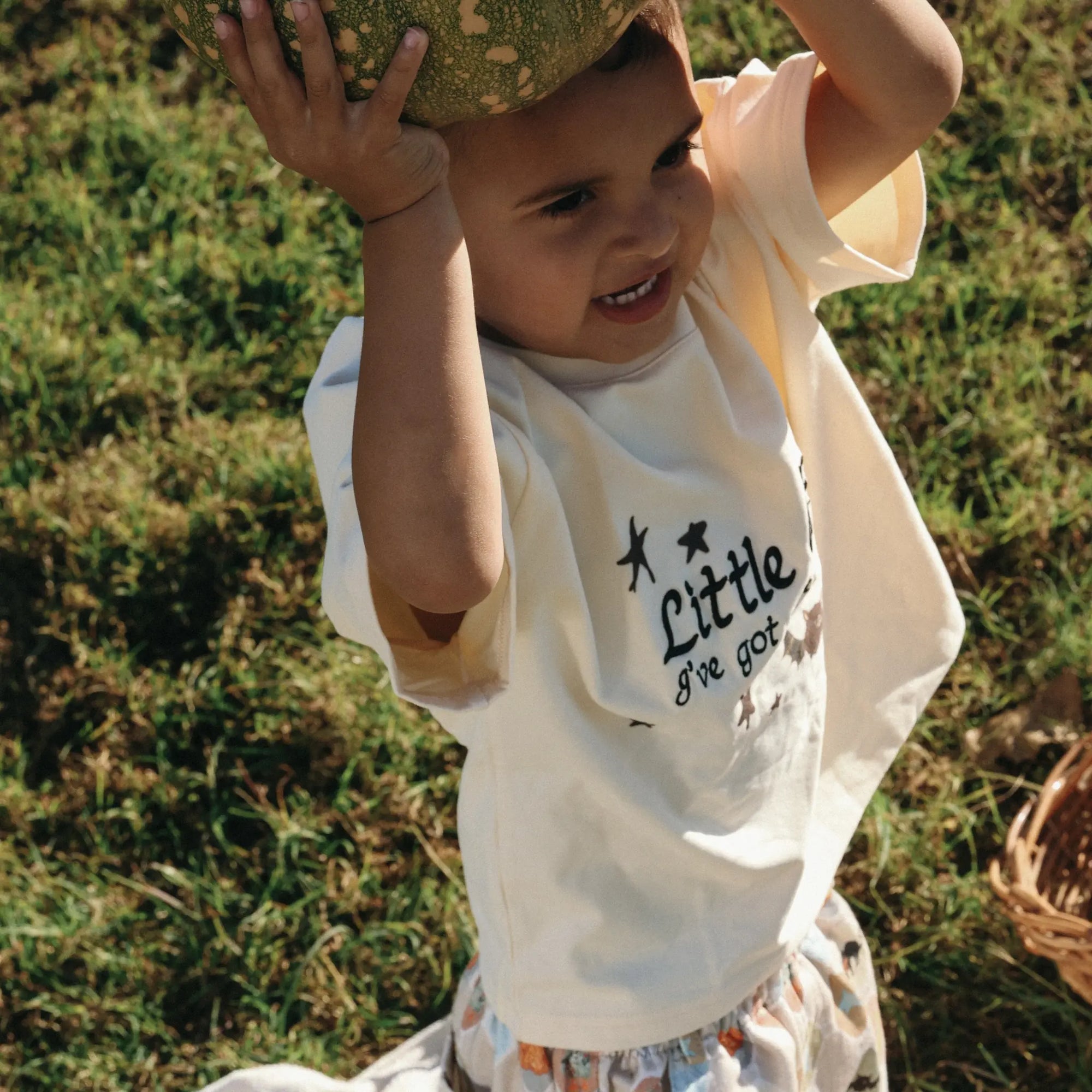 Toddler in a Little Charmer print tee balancing a pumpkin on his head in a sunlit field
