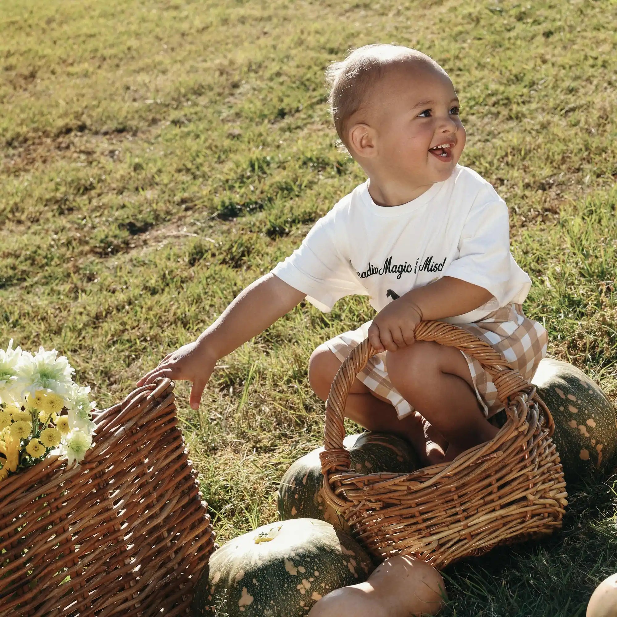 Baby in a Spreading Magic and Mischief tee and gingham shorts sitting between wicker baskets and pumpkins