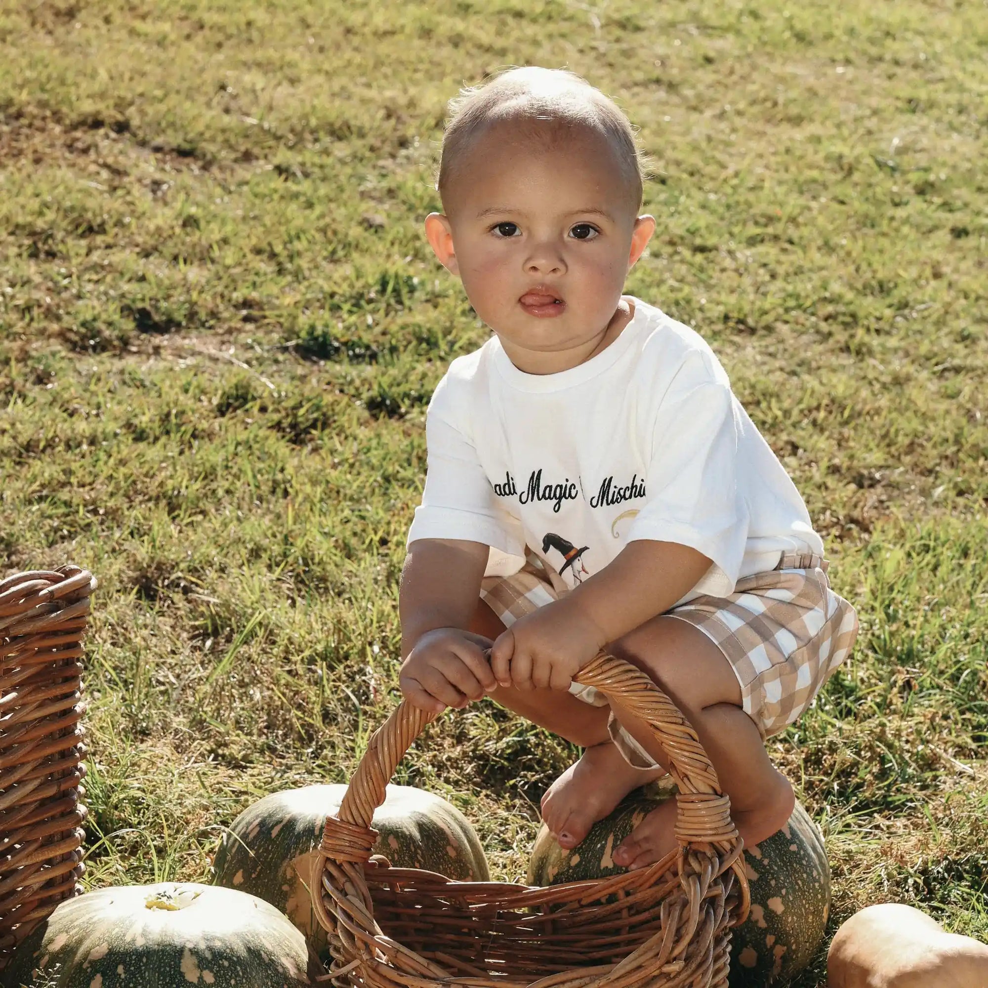 Baby in a Spreading Magic and Mischief tee and gingham shorts holding a wicker basket in a field