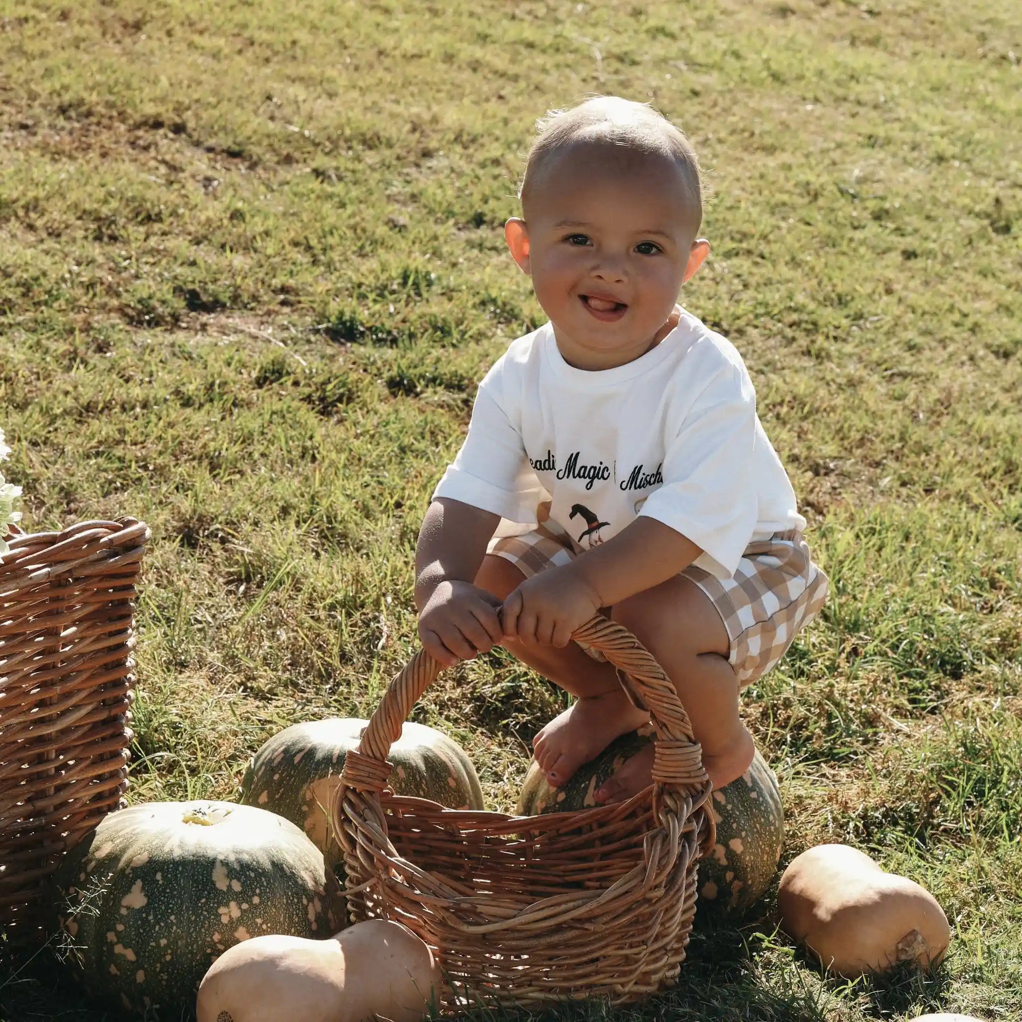 Smiling baby in a Spreading Magic and Mischief tee and tan gingham shorts crouching beside pumpkins