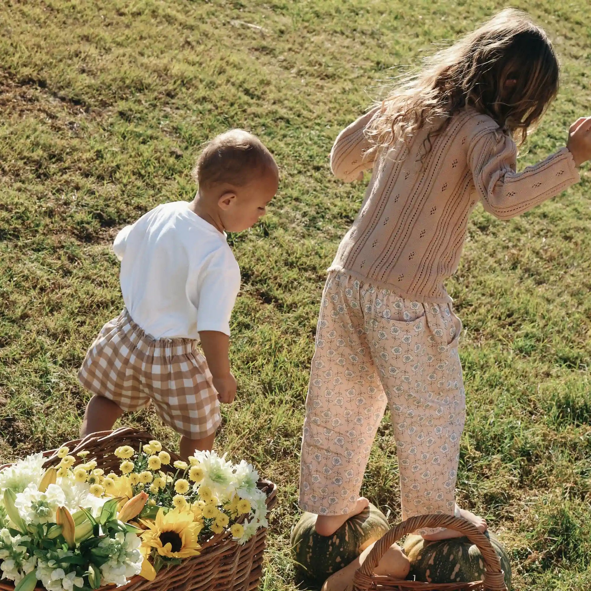 Toddler in a cream tee and tan gingham shorts beside a girl balancing on pumpkins in a field