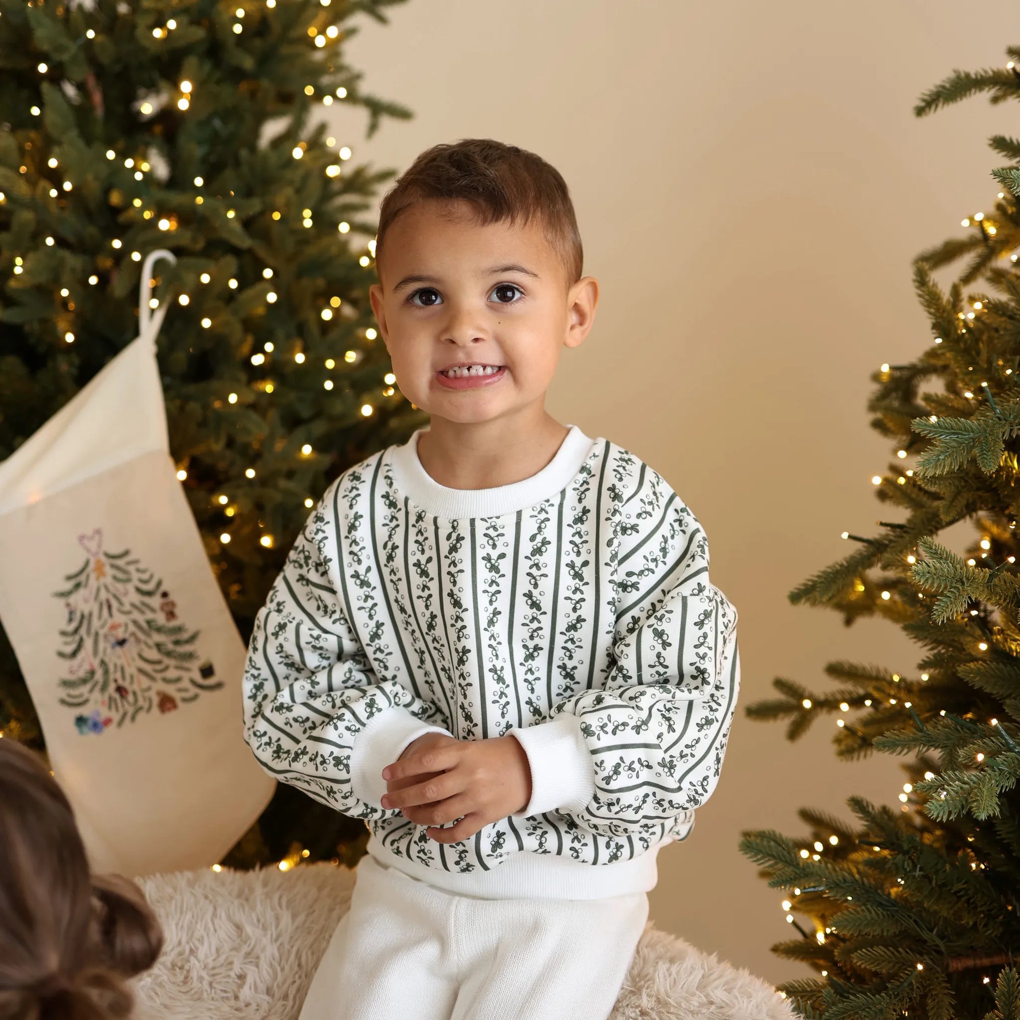 Boy in a cream knit Christmas jumper with a green botanical print and matching pants, beside an embroidered stocking