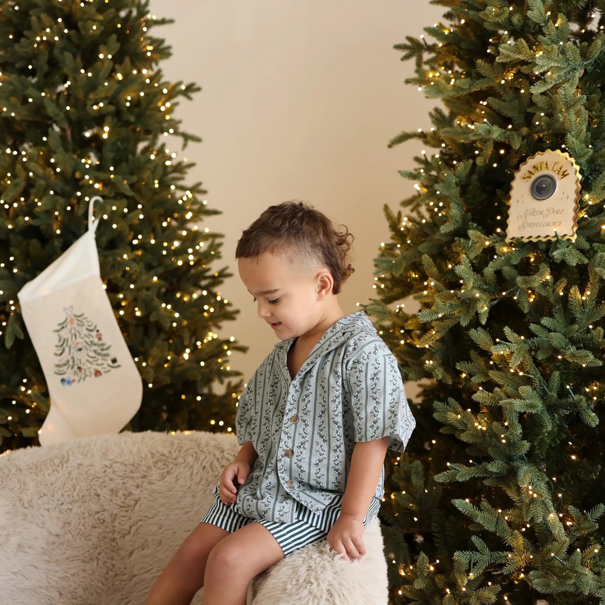 Toddler in a sage green botanical print shirt and stripe shorts sitting on a sheepskin chair between lit trees