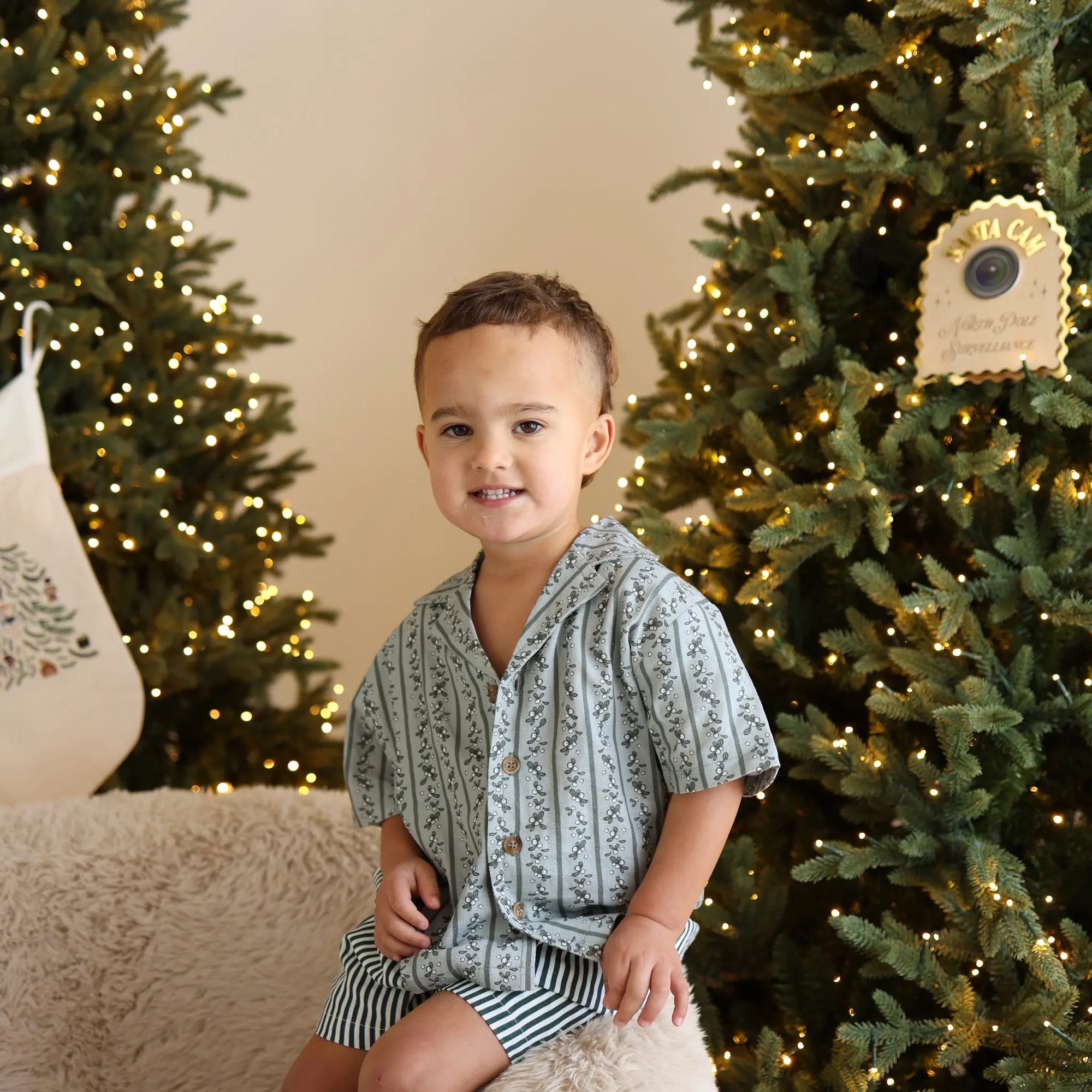 Toddler in a sage green botanical print pyjama shirt and stripe shorts beside a lit Christmas tree