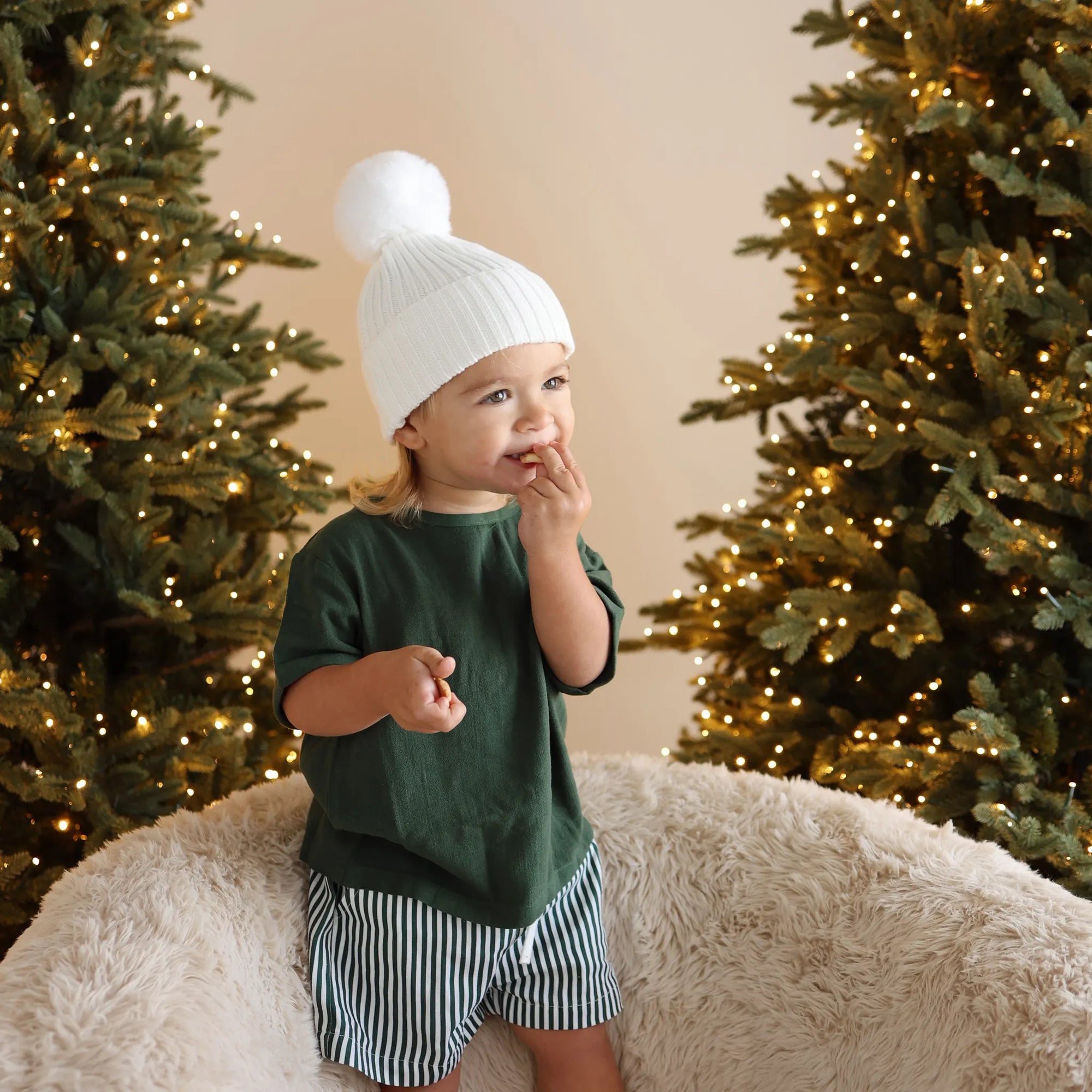 Toddler in a forest green tee, stripe shorts and a white pom beanie standing on a sheepskin chair