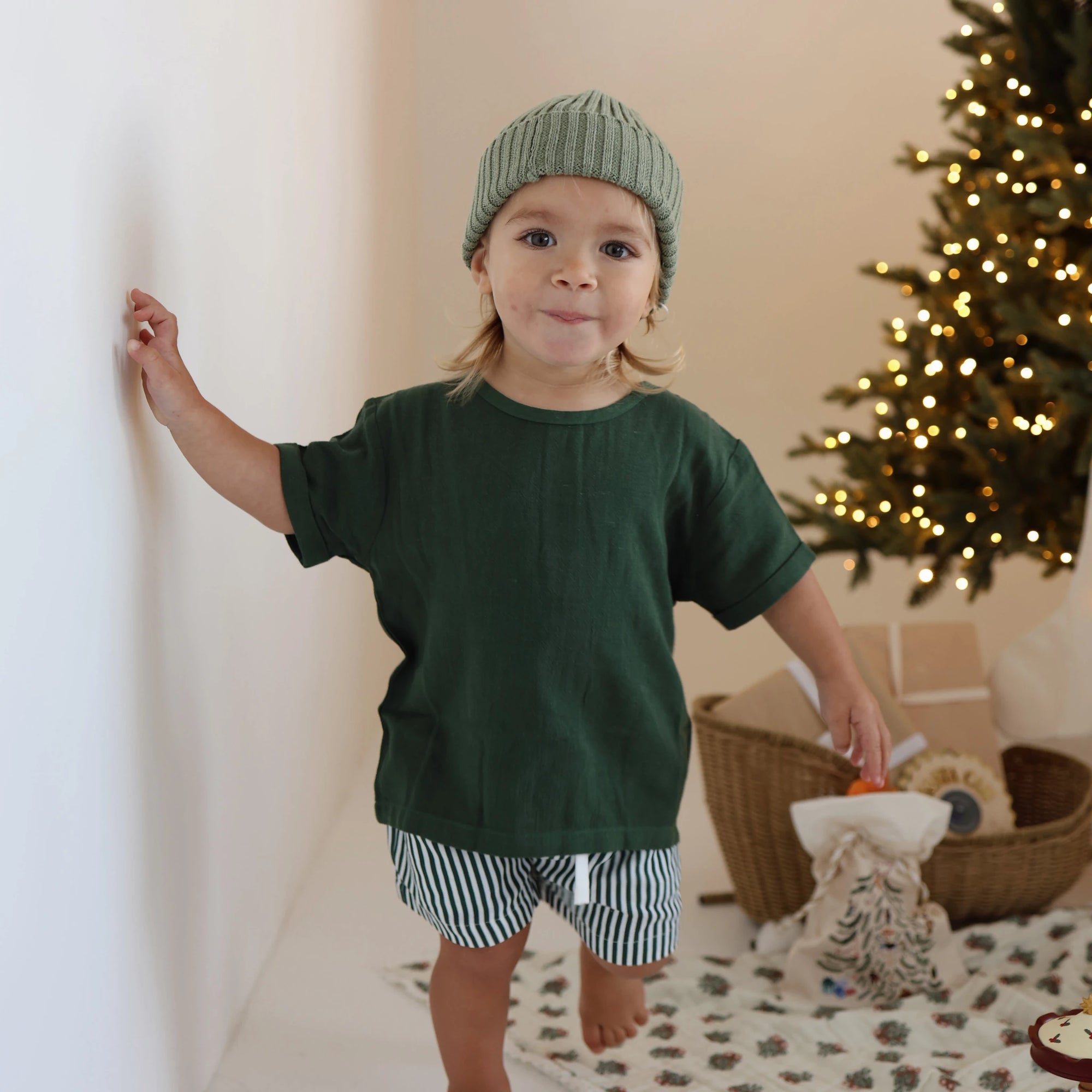 Toddler in a forest green tee, stripe shorts and a sage pom beanie beside a lit Christmas tree and gift basket
