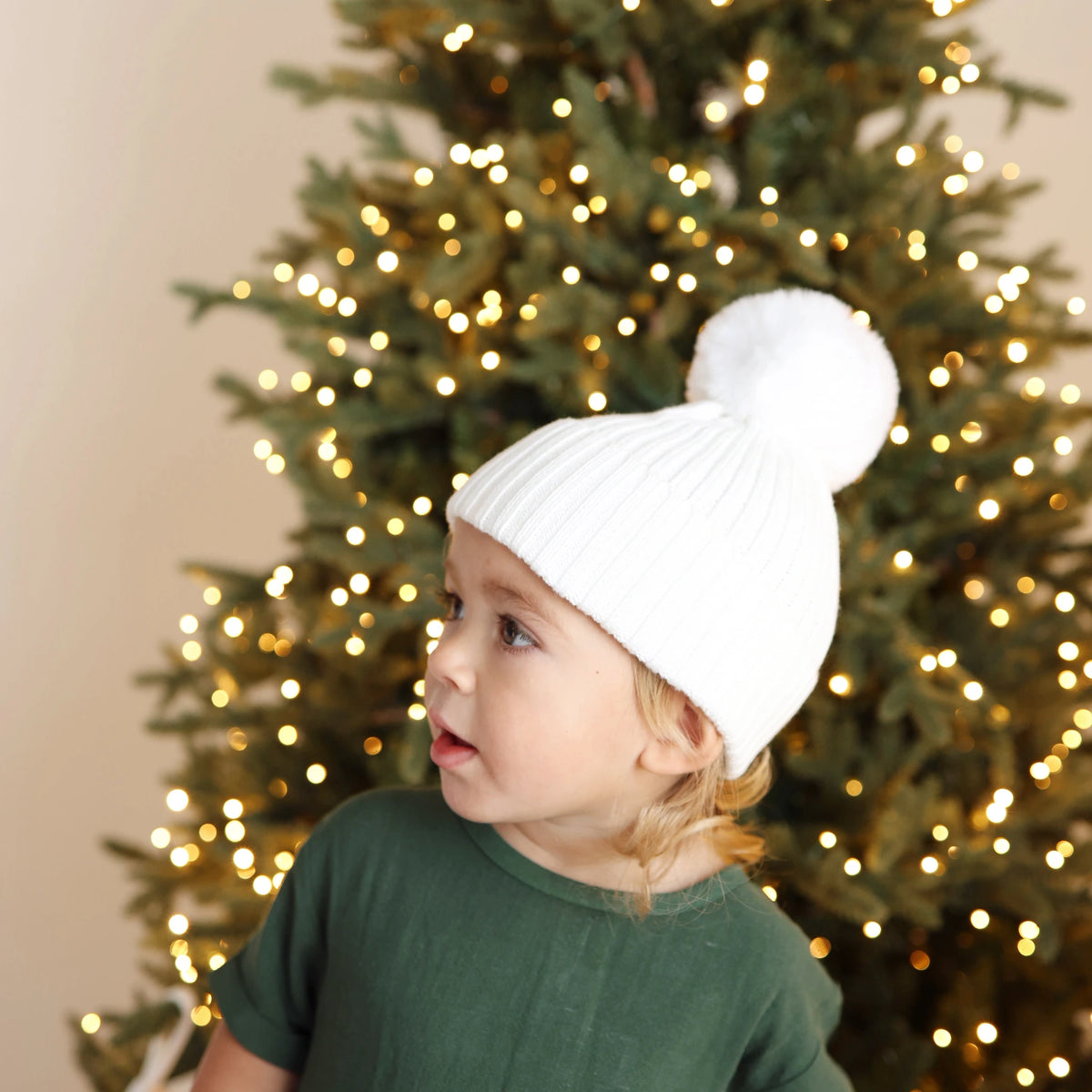 Toddler in a forest green tee and white pom beanie in front of a lit Christmas tree