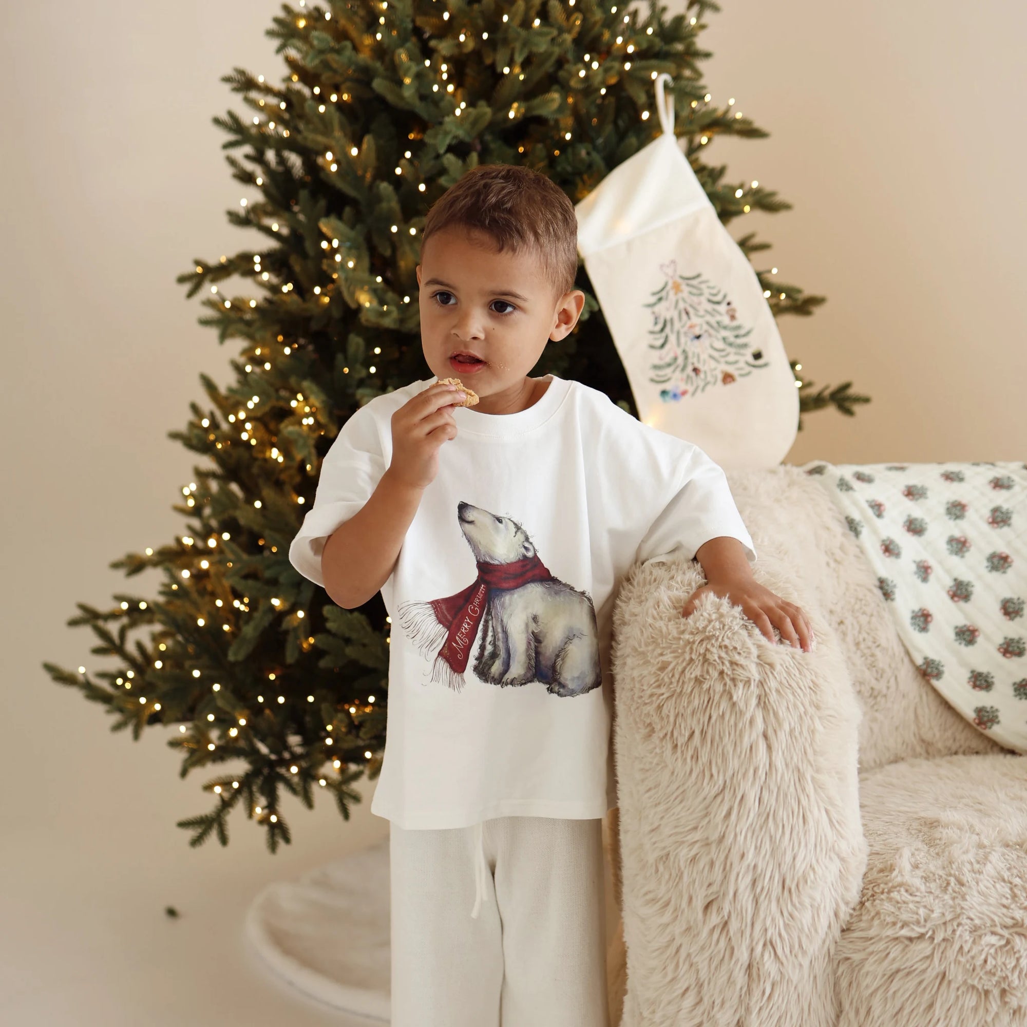 Boy in a polar bear print tee and cream pants eating a cookie beside a lit Christmas tree and stocking