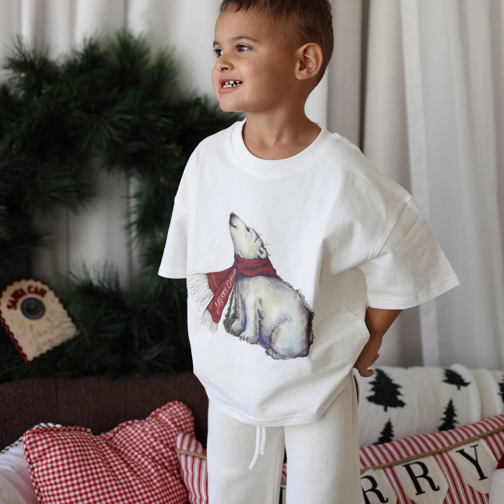Boy in a polar bear print tee and cream pants beside a Christmas wreath and festive cushions