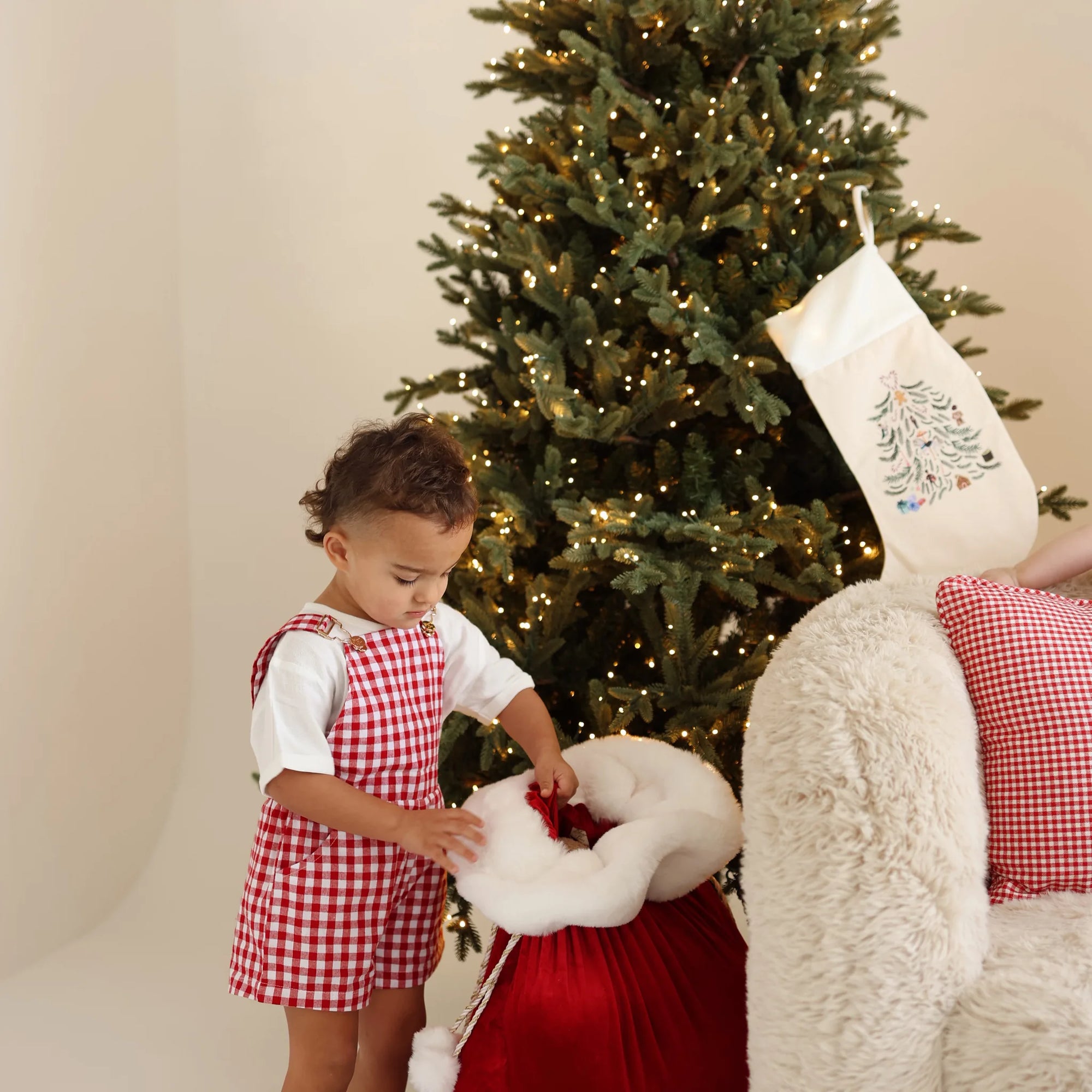 Toddler in a red gingham overall set opening a red velvet Santa sack beside a lit Christmas tree