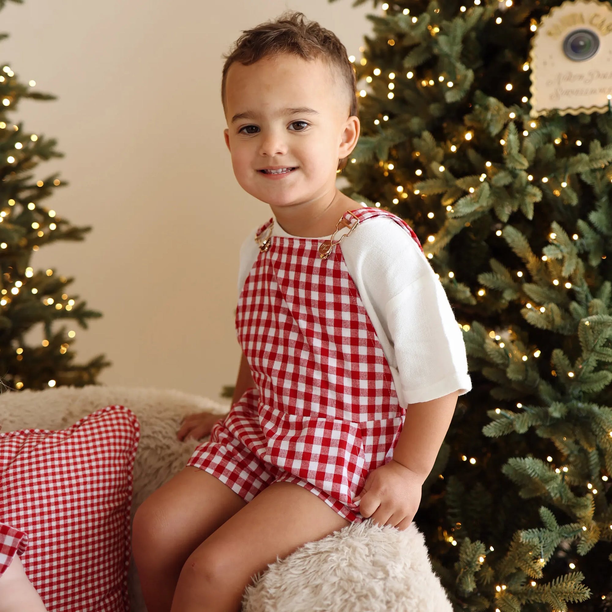 Toddler in a red gingham overall set and white tee sitting on a sheepskin chair by a lit tree
