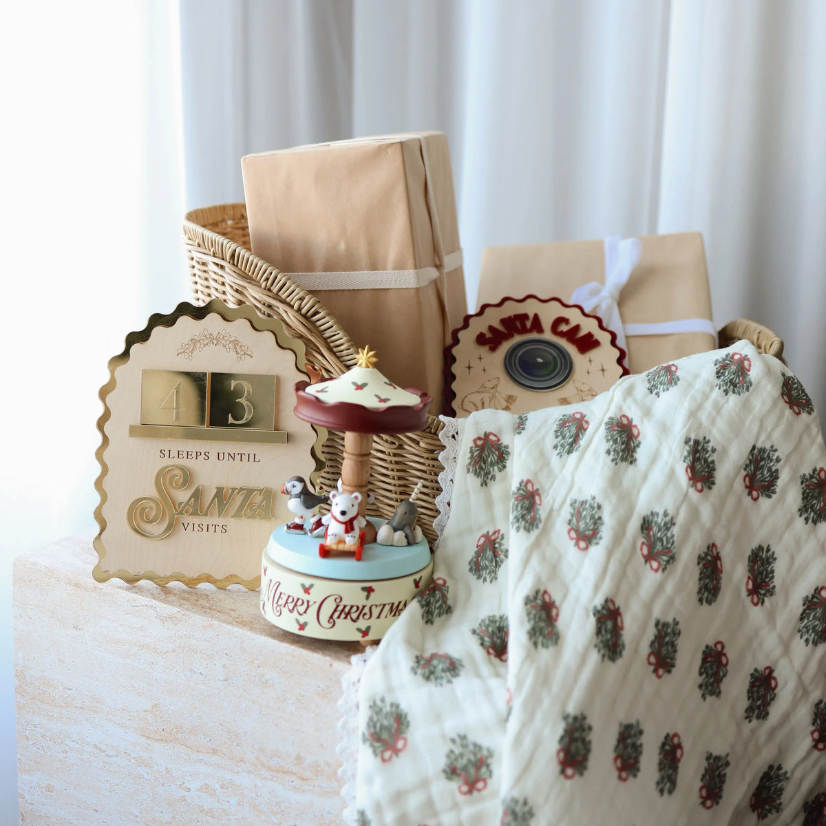 A styled holiday gift basket arrangement on a light wood surface with white curtains in the background. The basket contains kraft paper gift boxes, a wicker bag, a decorative wooden sign reading 'SLEEPS UNTIL SANTA VISITS', a small Christmas-themed bird music box, and a cream quilted blanket with a red and green holly pattern.