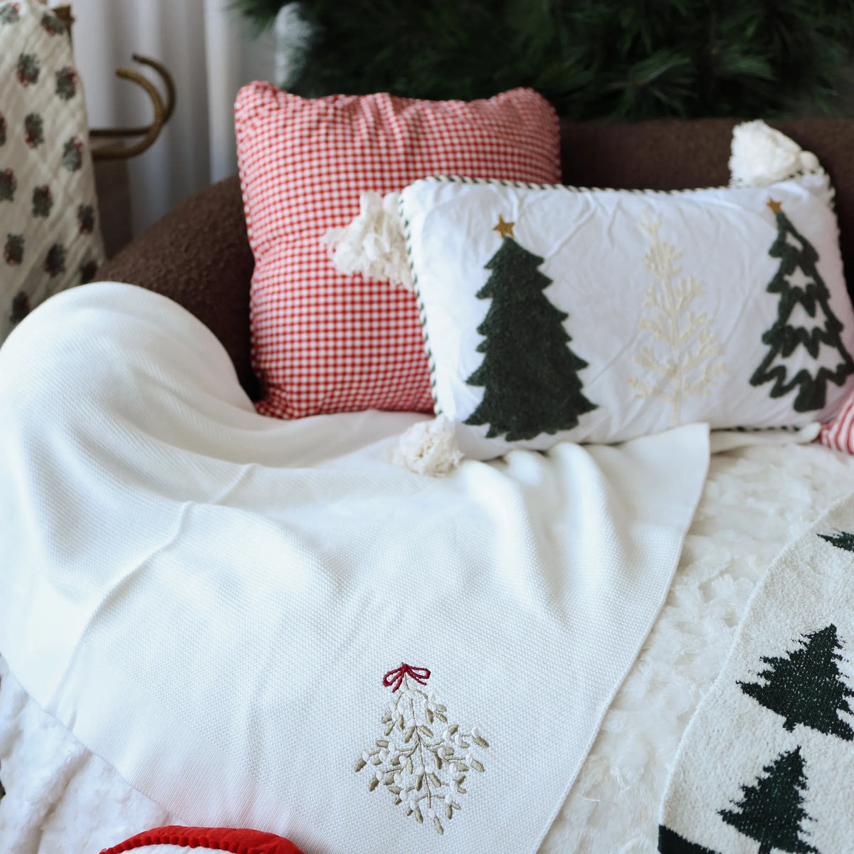 Christmas cushions and a mistletoe embroidered knit throw styled on a sofa, including a red gingham cushion and an embroidered tree cushion