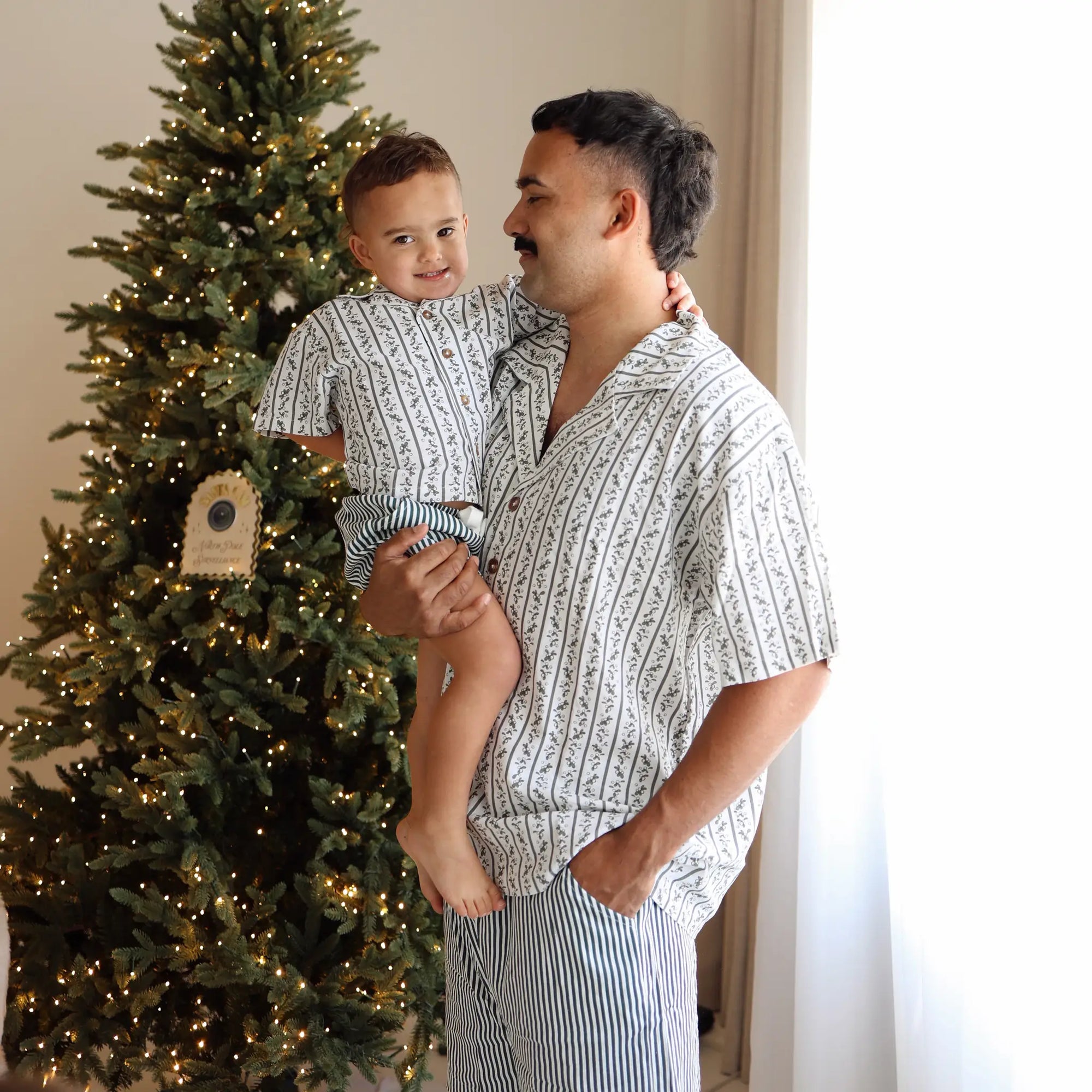 Dad holding a toddler in matching green botanical print shirts and stripe pyjama pants beside a lit tree