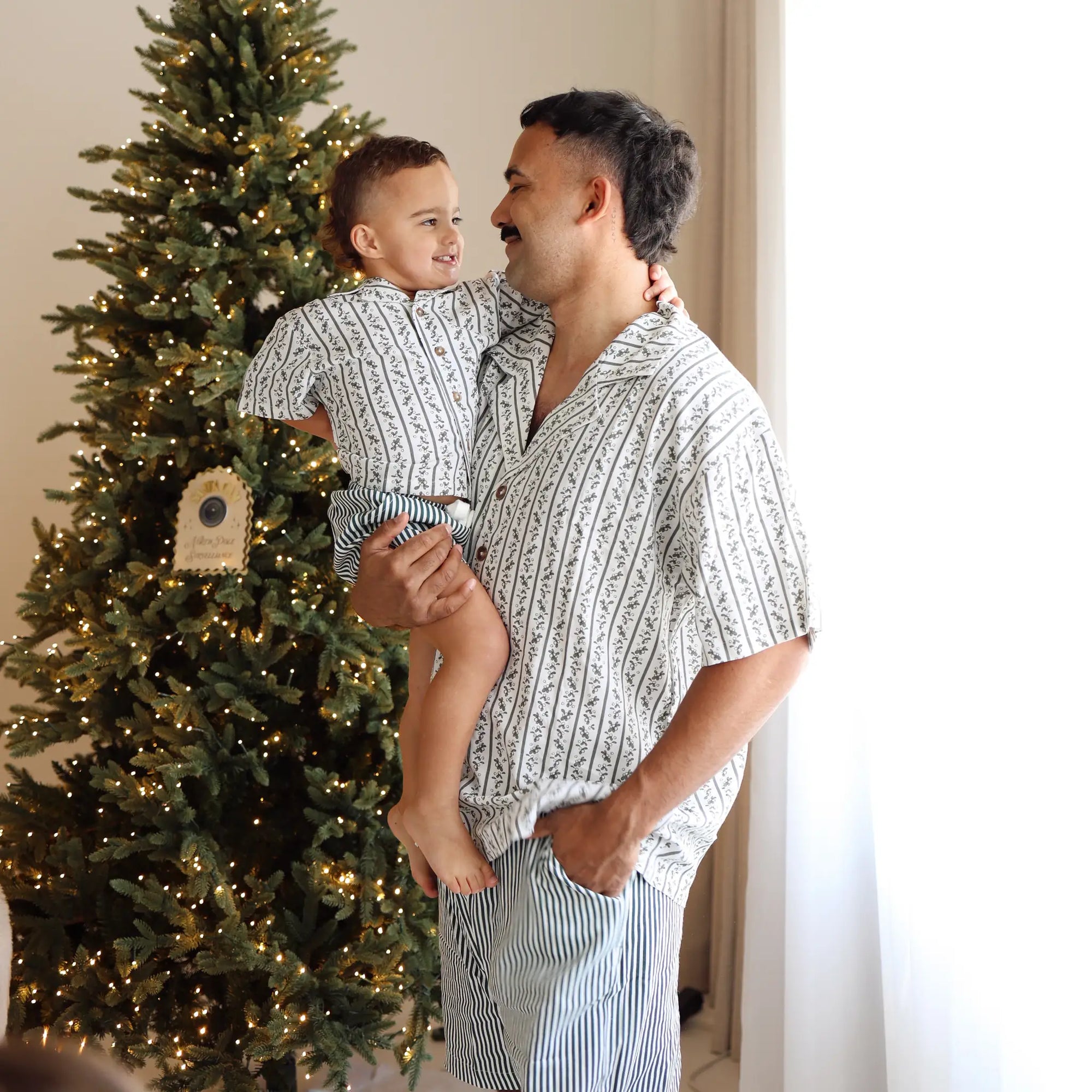 Dad and toddler smiling at each other in matching botanical print Christmas pyjamas beside a lit tree