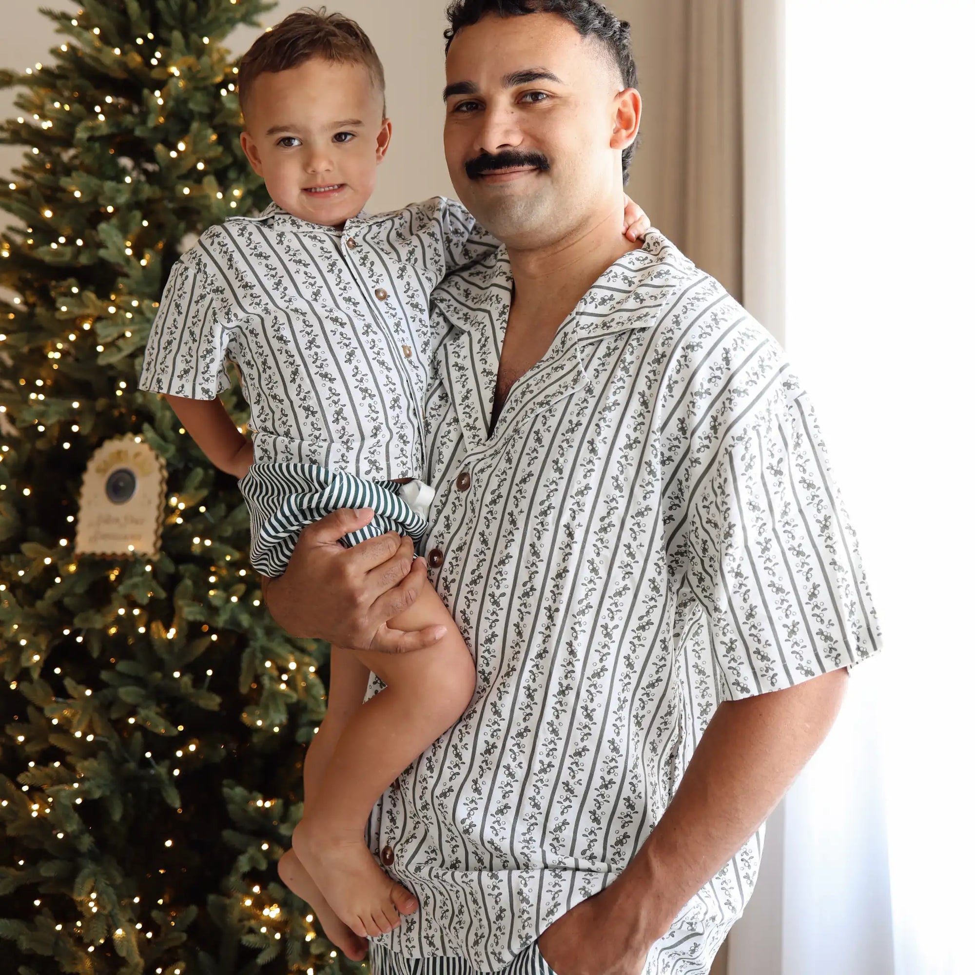 Dad holding a toddler in matching botanical print Christmas pyjama shirts beside a lit tree