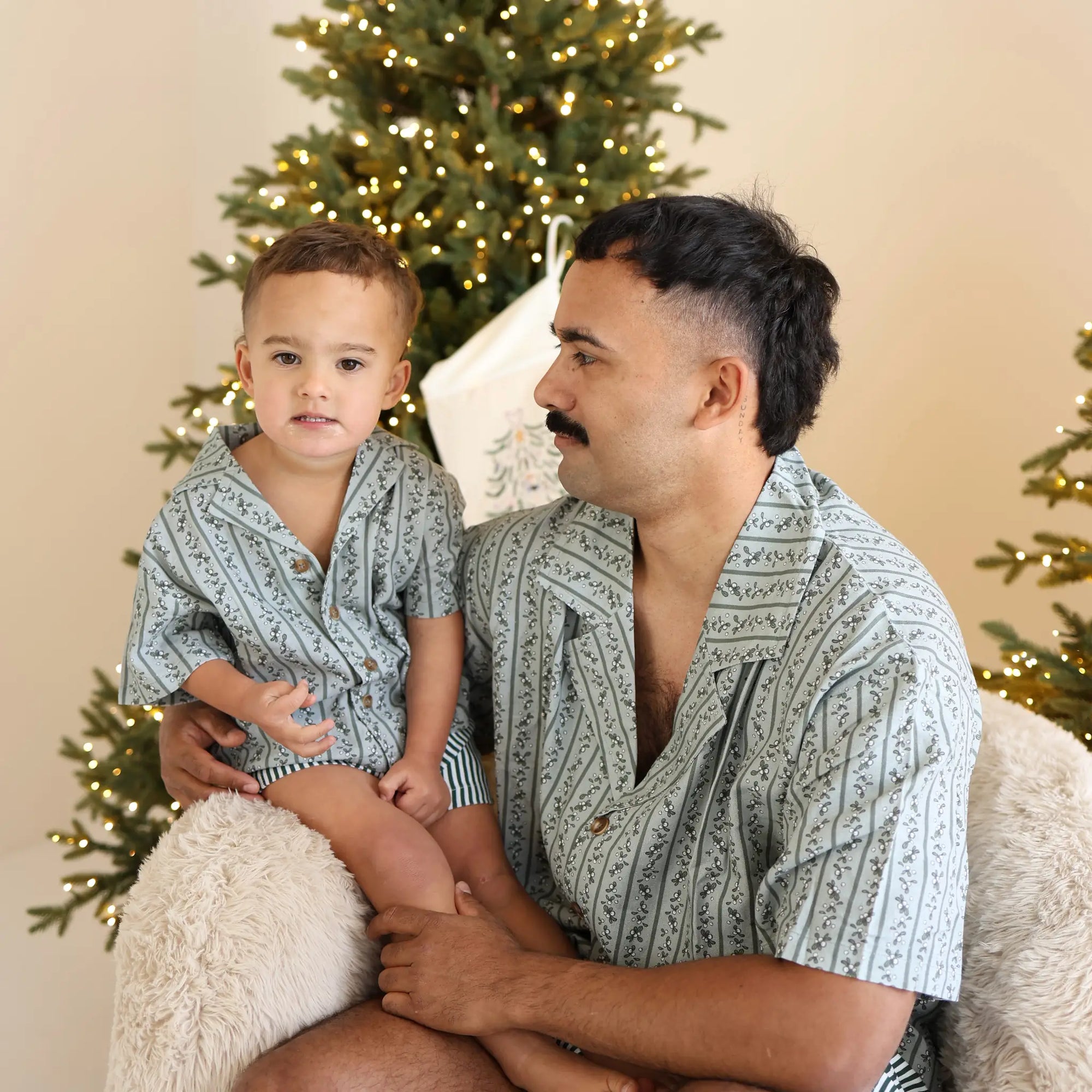 Dad holding a toddler on a sheepskin chair, both in matching sage green botanical print pyjama shirts