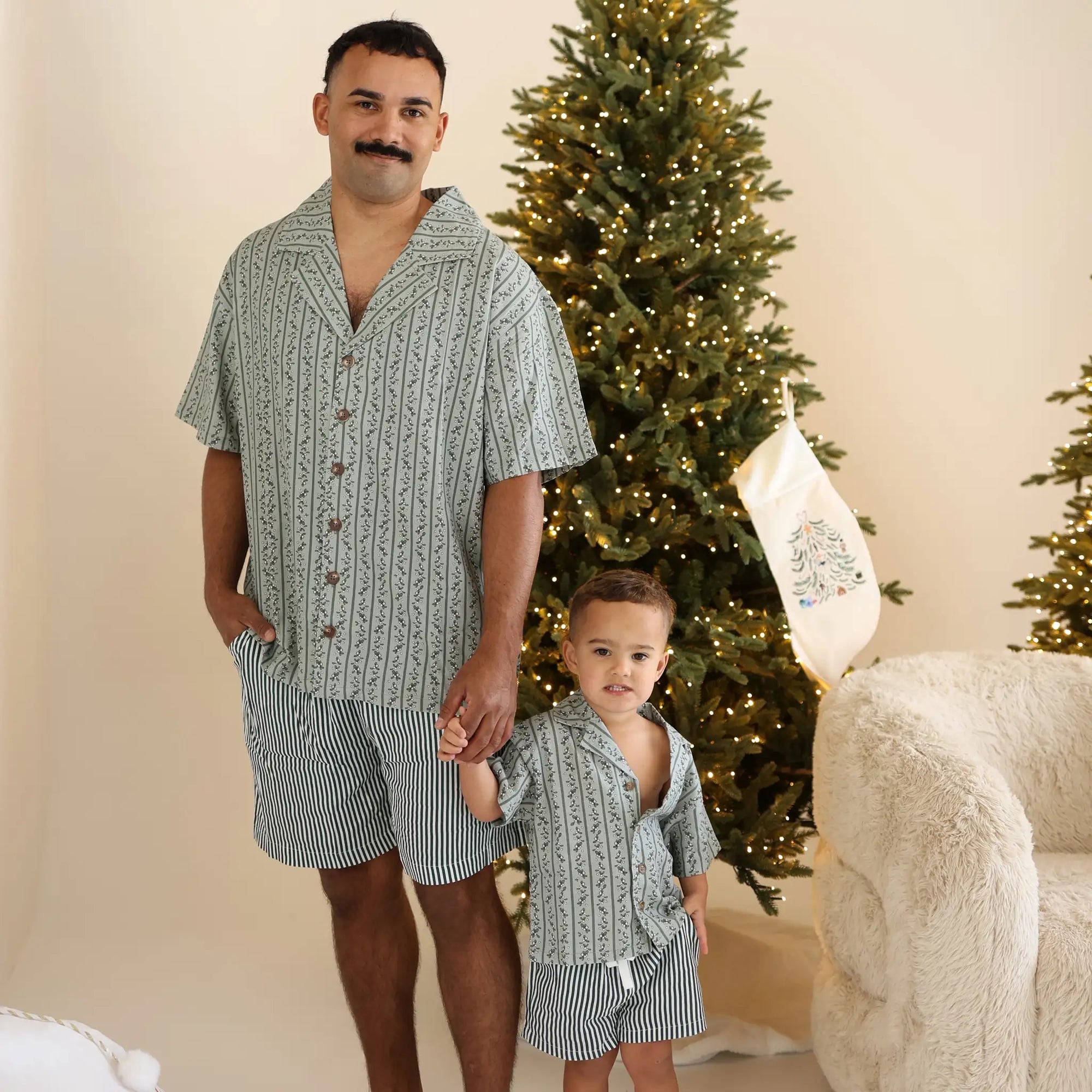 Dad and toddler holding hands in matching green botanical print shirts and stripe shorts beside a lit Christmas tree