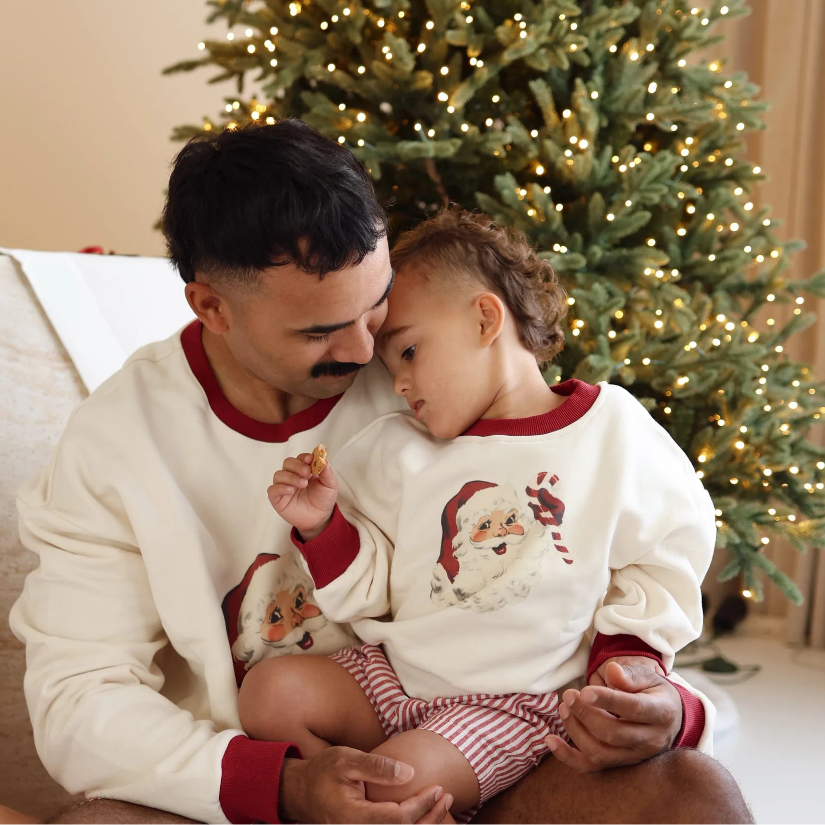 Dad and toddler in matching vintage Santa print jumpers sharing a quiet moment by a lit Christmas tree