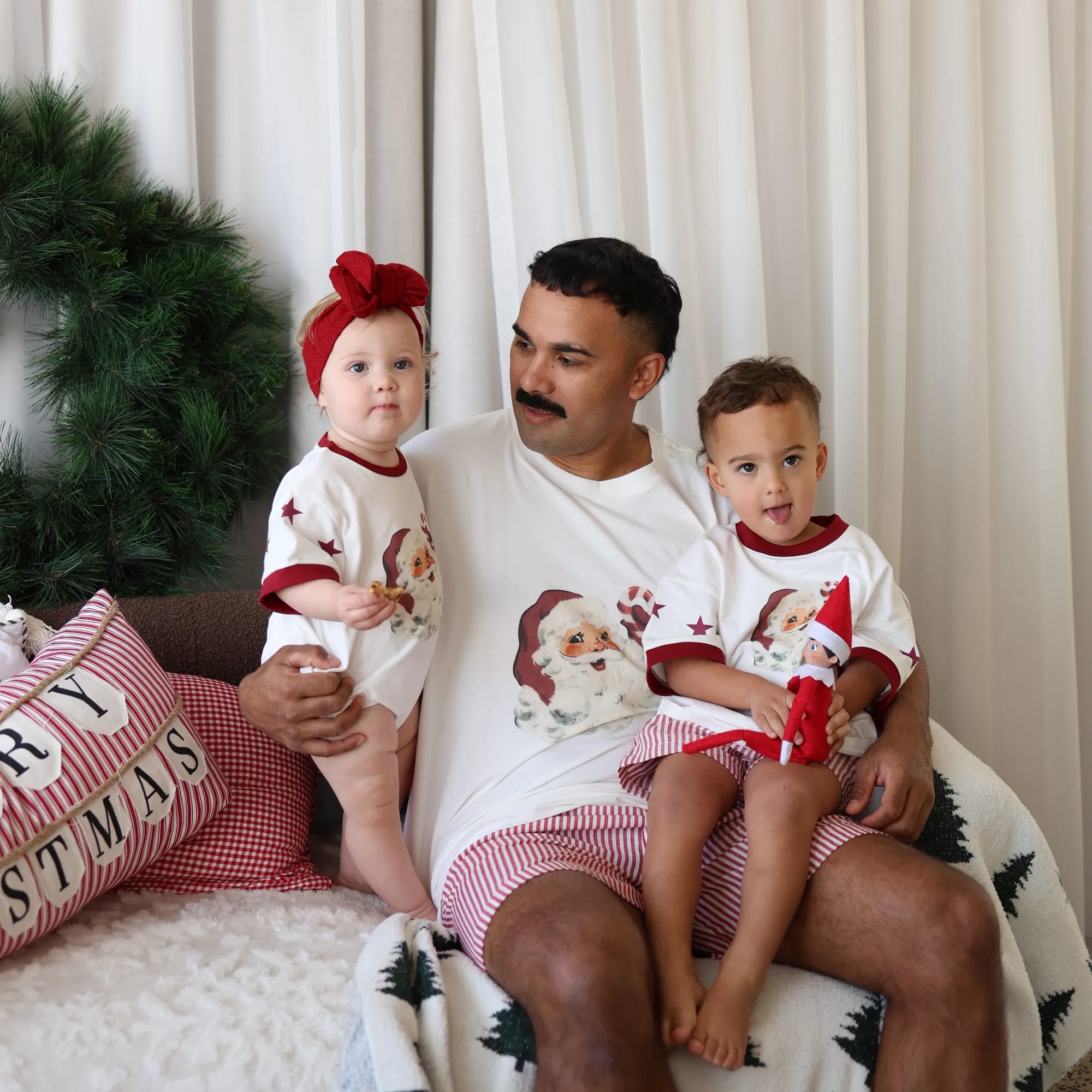 Dad and two children in matching Santa print pyjamas on a sofa, one holding an elf doll, with Merry Christmas cushions
