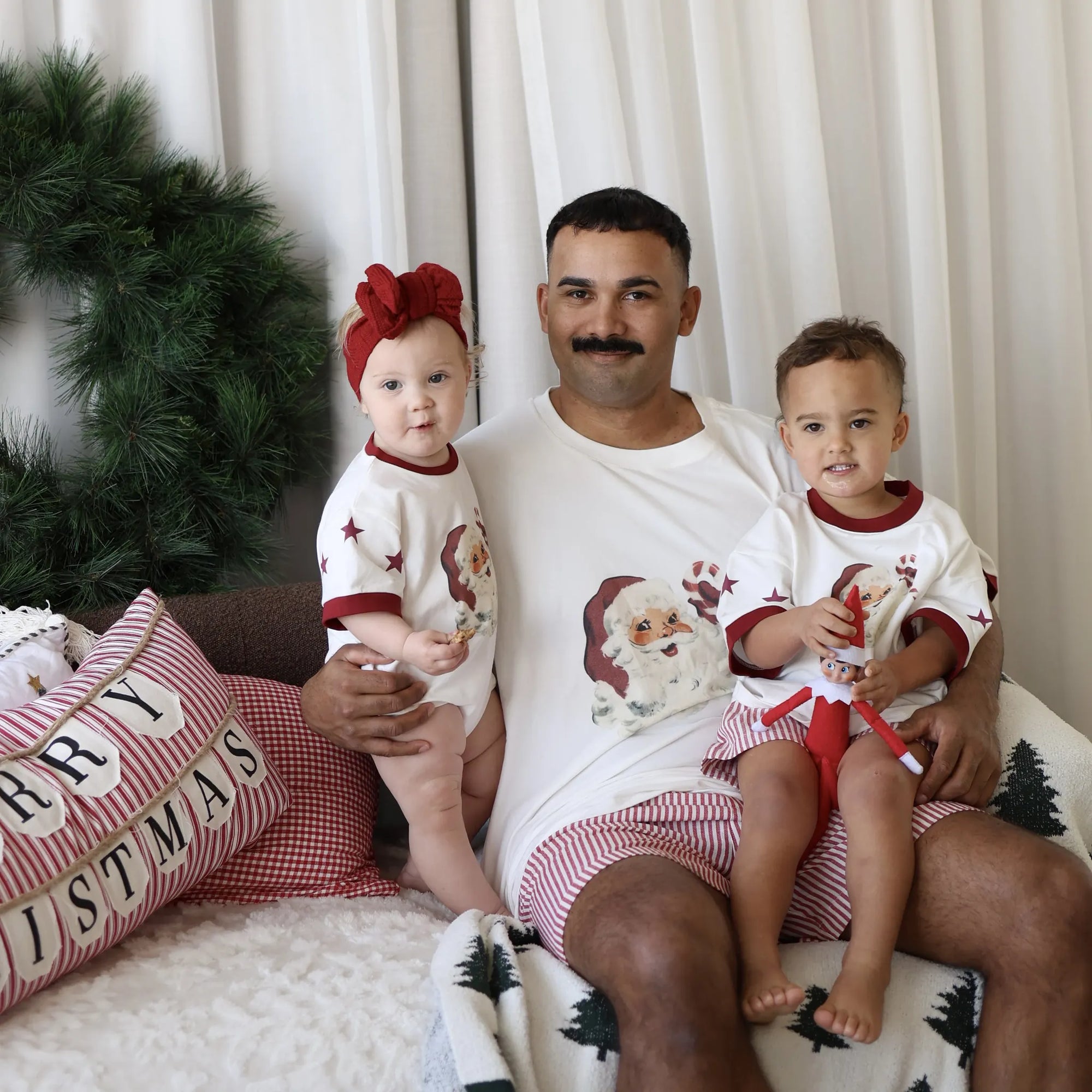 Dad holding two children in matching vintage Santa print pyjamas on a sofa with Merry Christmas cushions