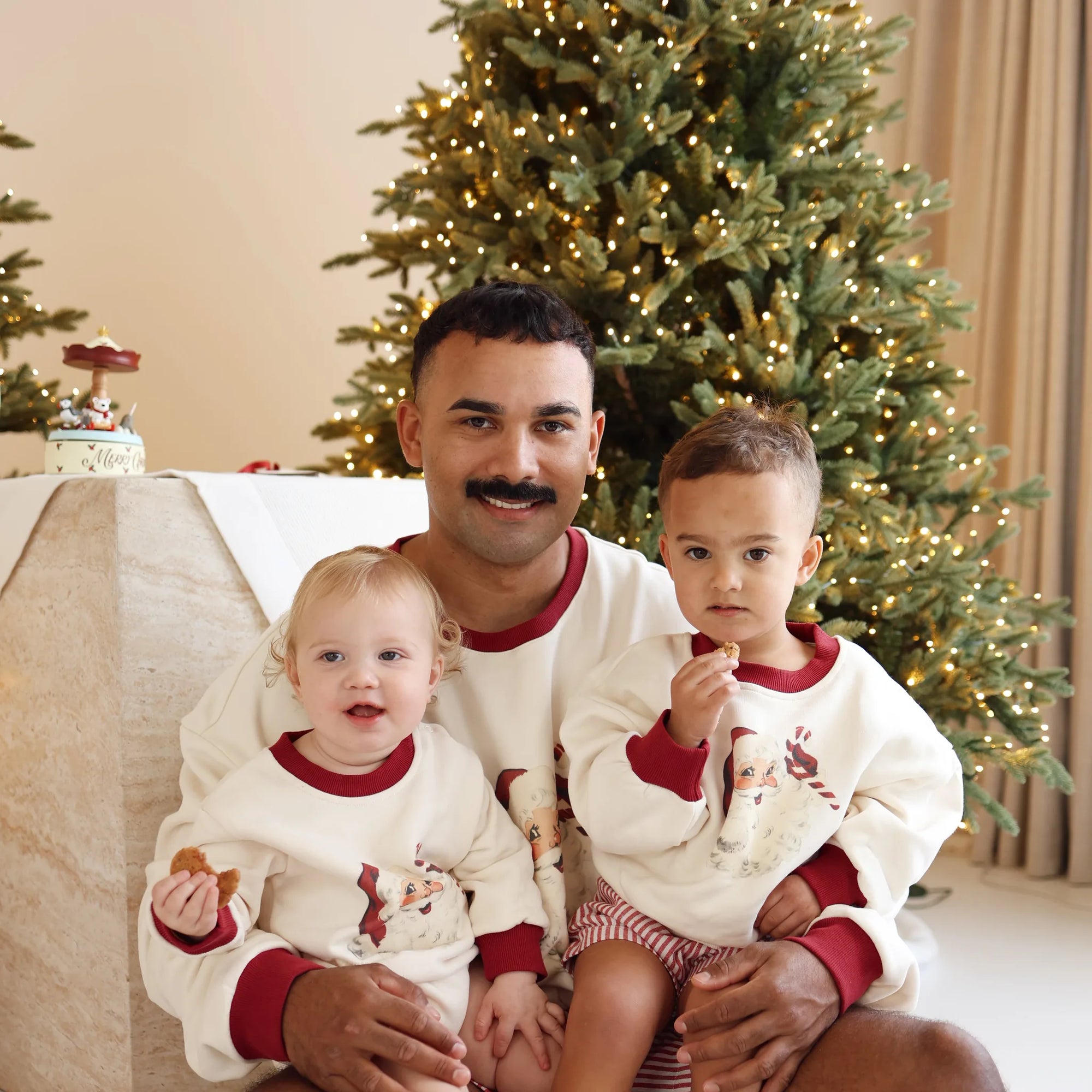 Dad holding two toddlers, all in matching vintage Santa print jumpers, in front of a lit Christmas tree