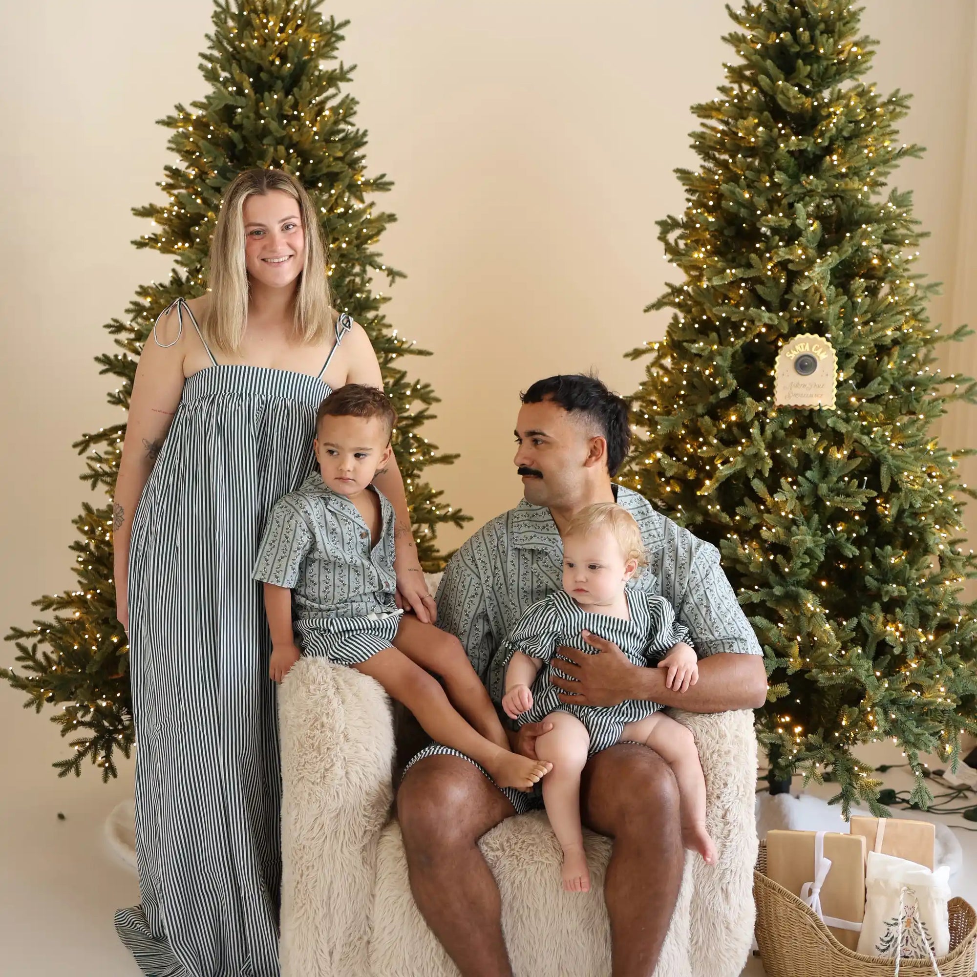 Family of four in matching green stripe and botanical print Christmas pyjamas between lit trees